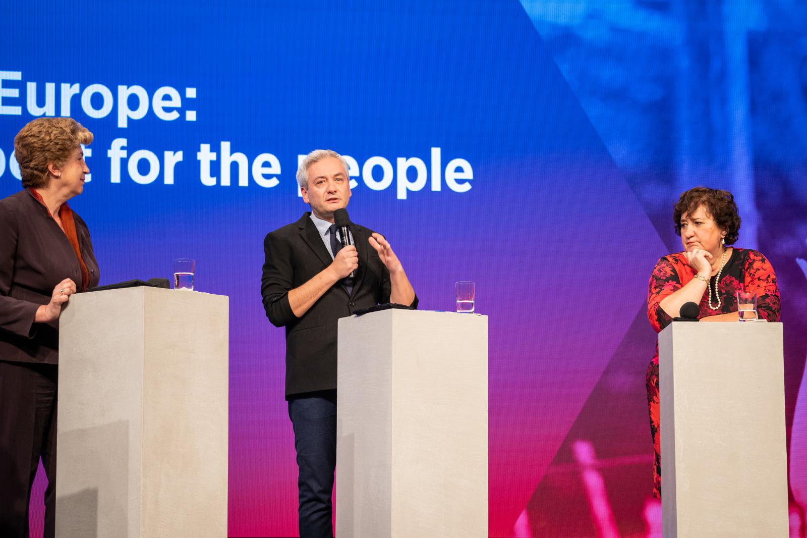 Na zdjęciu widać trzech uczestników debaty, z mikrofonem i notatkami, prowadzących dyskusję na tle kolorowego tła z napisem