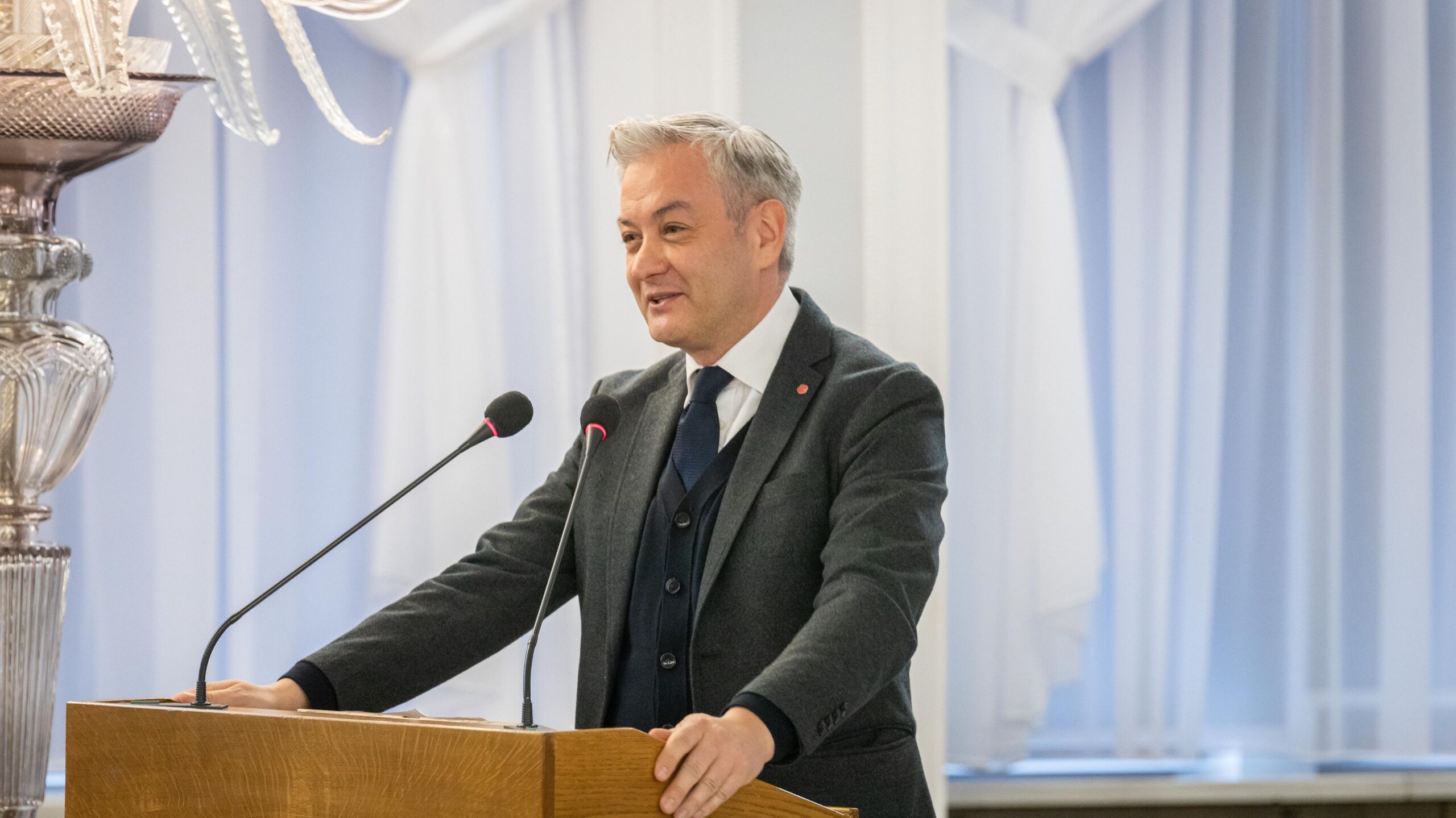 Mężczyzna w garniturze stoi za drewnianym pulpitem z dwoma mikrofonami i wygłasza przemówienie podczas konferencji, a w tle widać jasne zasłony.