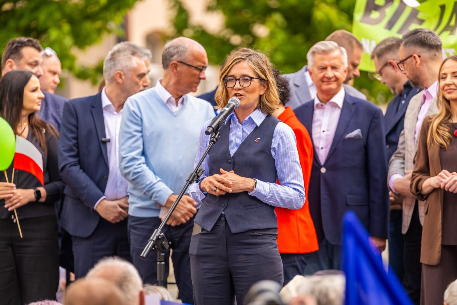 Kobieta w okularach, granatowej kamizelce i koszuli przemawia do mikrofonu podczas Święta Pracy 2025, otoczona grupą osób w garniturach.