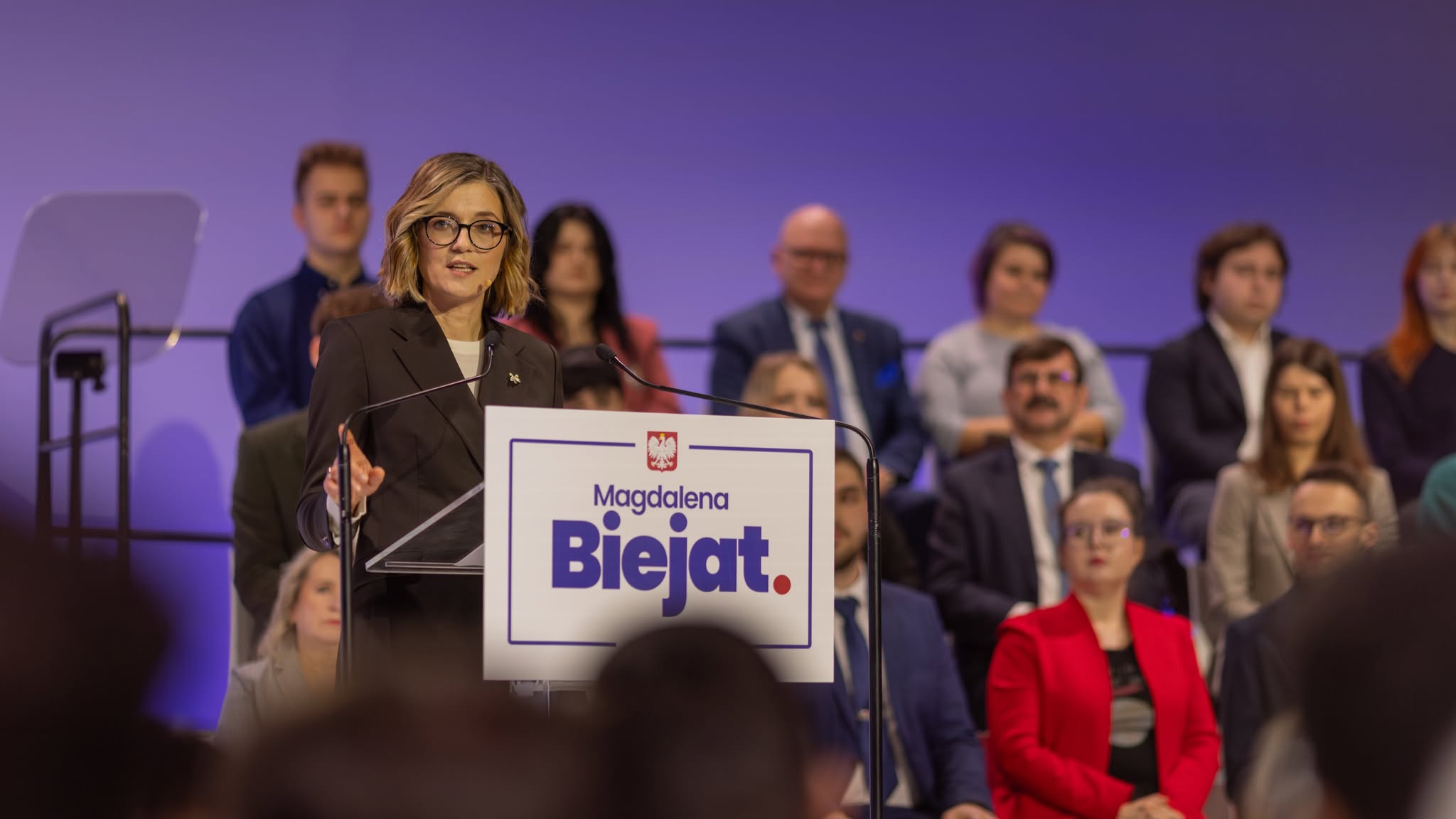 Kobieta z okularami wygłasza przemówienie z podium z napisem „Magdalena Biejat” podczas wydarzenia, na tle zgromadzonej widowni.