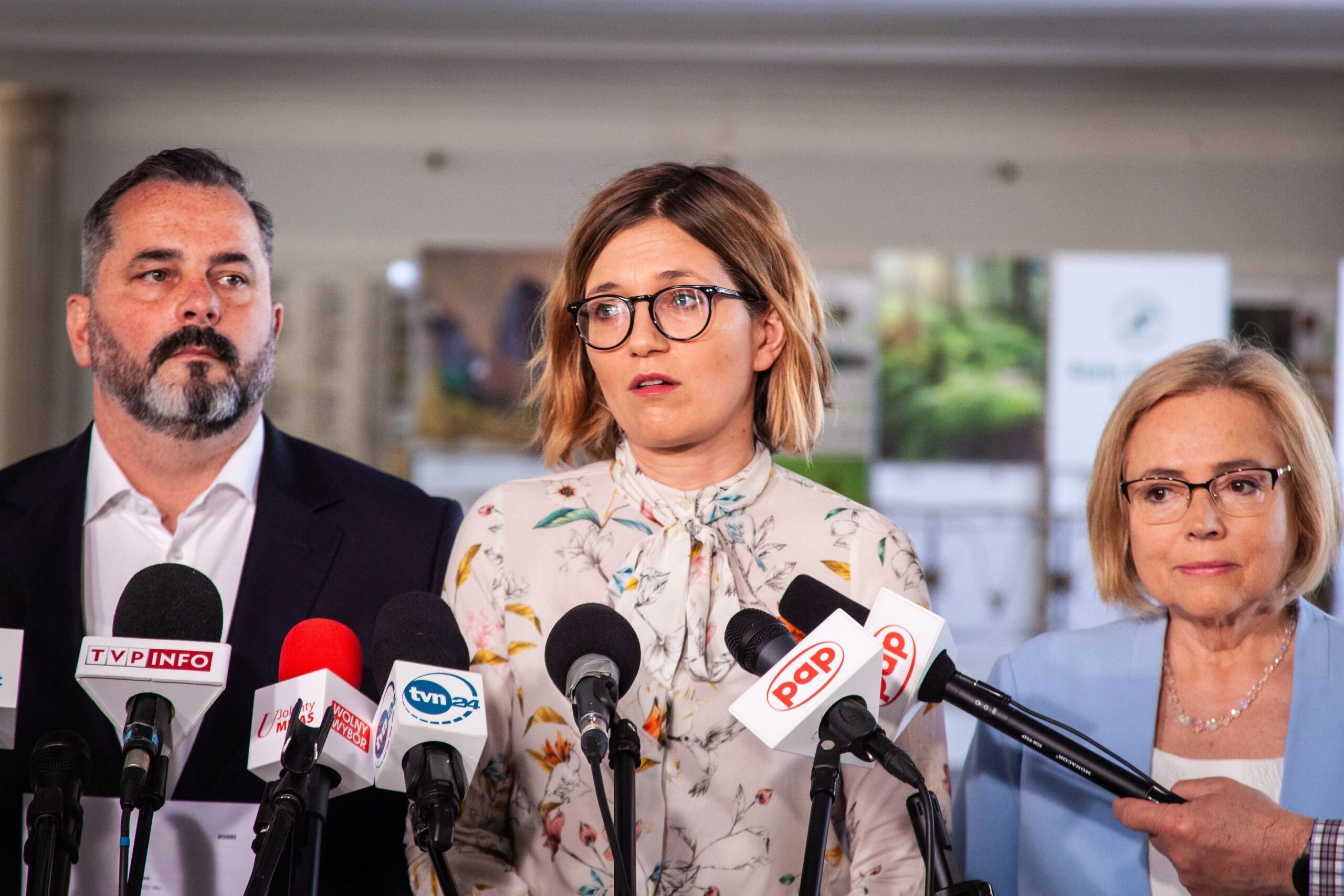 Trzy osoby stoją na konferencji prasowej dotyczącej pakietu Bezpieczny Senior; pośrodku kobieta w okularach przemawia do licznych mikrofonów, obok niej mężczyzna i starsza kobieta.