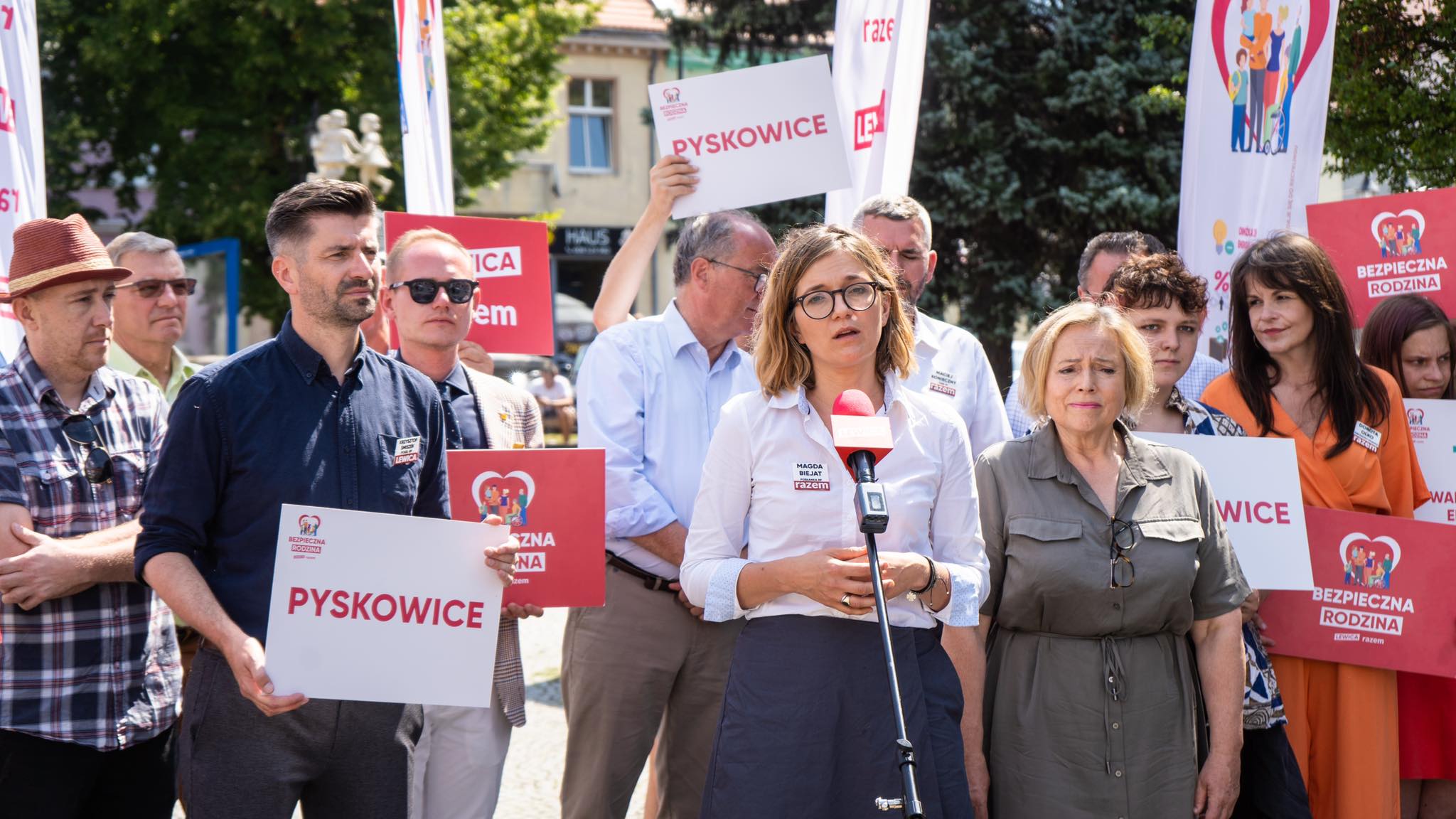 Kobieta przemawia do mikrofonu podczas wiecu Lewicy w Pyskowicach, w tle grupa ludzi trzyma plakaty z napisem Pyskowice i loga partii.