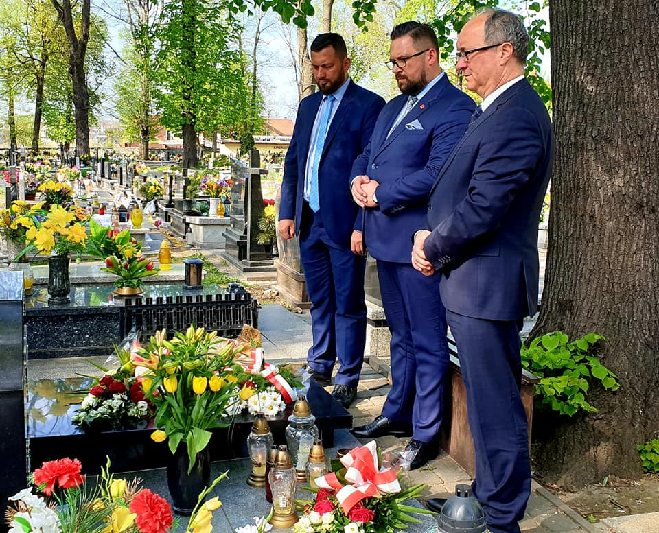 Trzech mężczyzn w garniturach stoi przy nagrobku na cmentarzu, w otoczeniu kwiatów i zniczy.