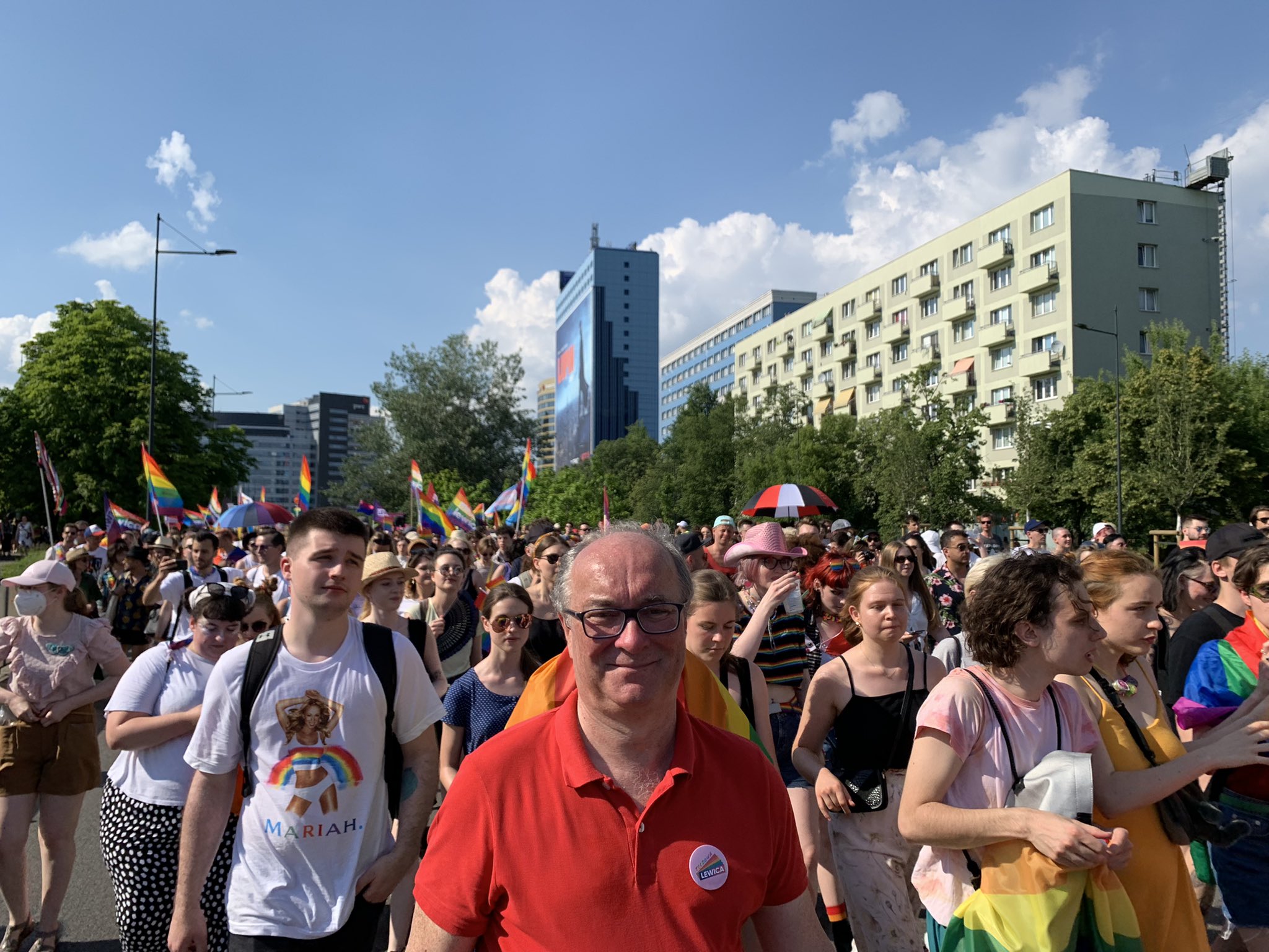 Duży tłum uczestników Parady Równości 2021 przechodzi ulicami miasta, trzymając flagi w kolorach tęczy i ubrania w barwach solidarności, w słoneczny dzień.