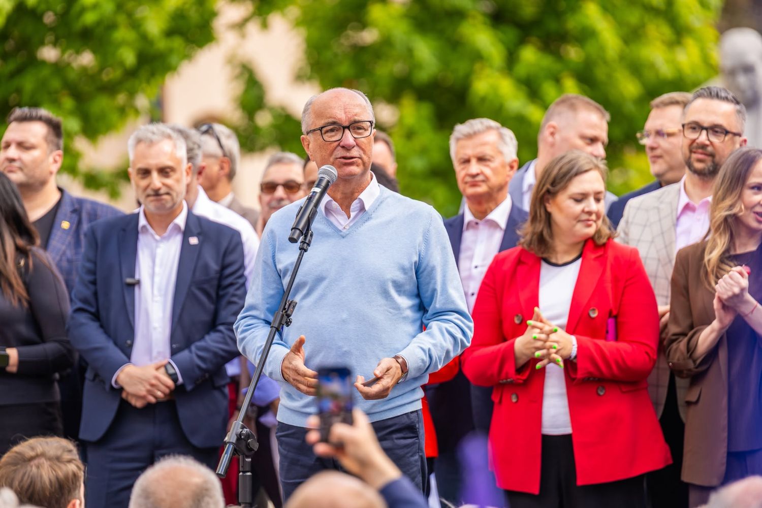 Mężczyzna w jasnoniebieskim swetrze przemawia do mikrofonu podczas Święta Pracy 2025, otoczony grupą uczestników w garniturach i marynarkach.