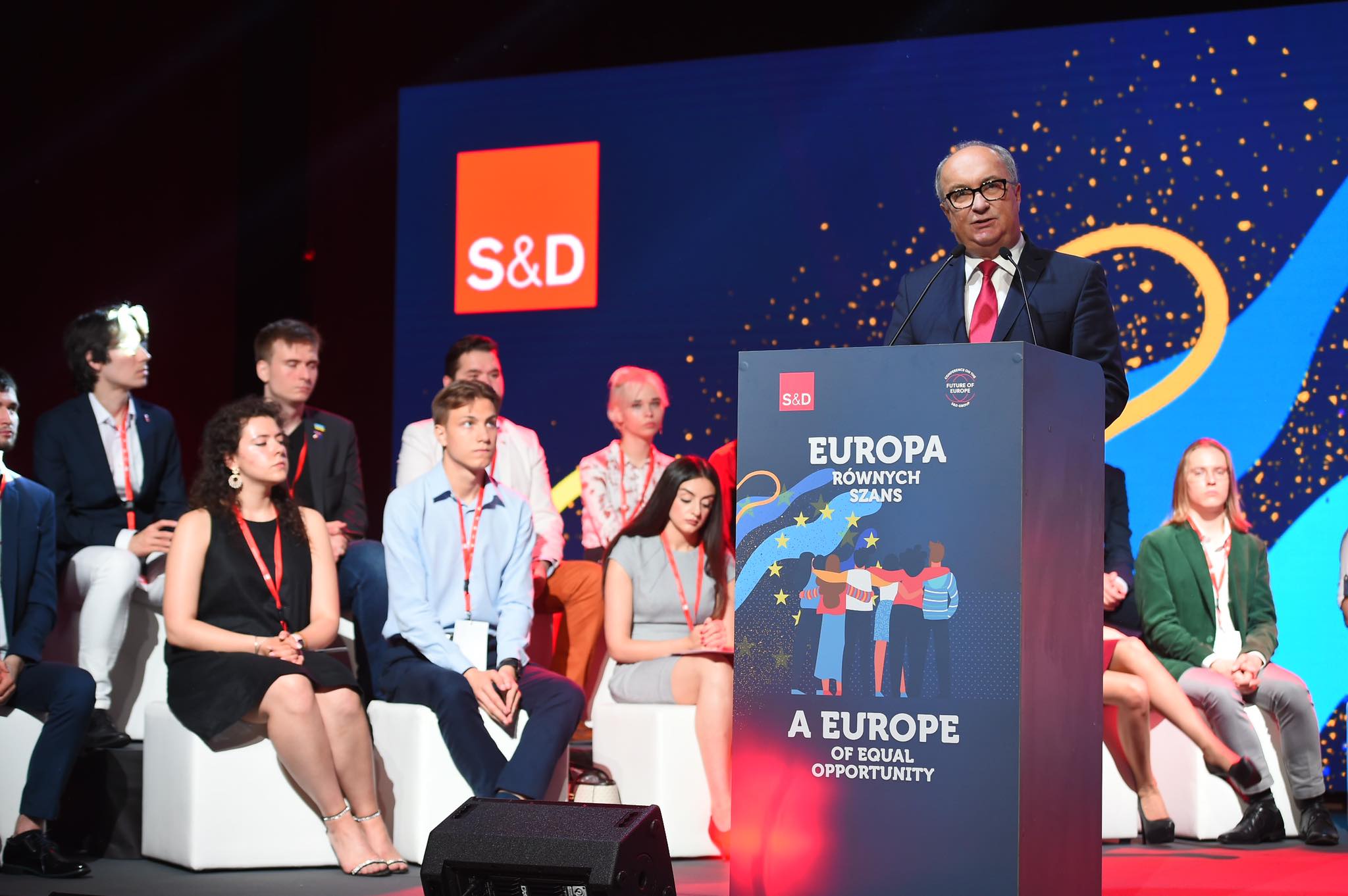 Na zdjęciu widać wystąpienie mężczyzny przy mównicy, za którym siedzi grupa młodych ludzi, a w tle znajdują się grafiki przedstawiające hasła związane z równością i europejskimi wartościami.