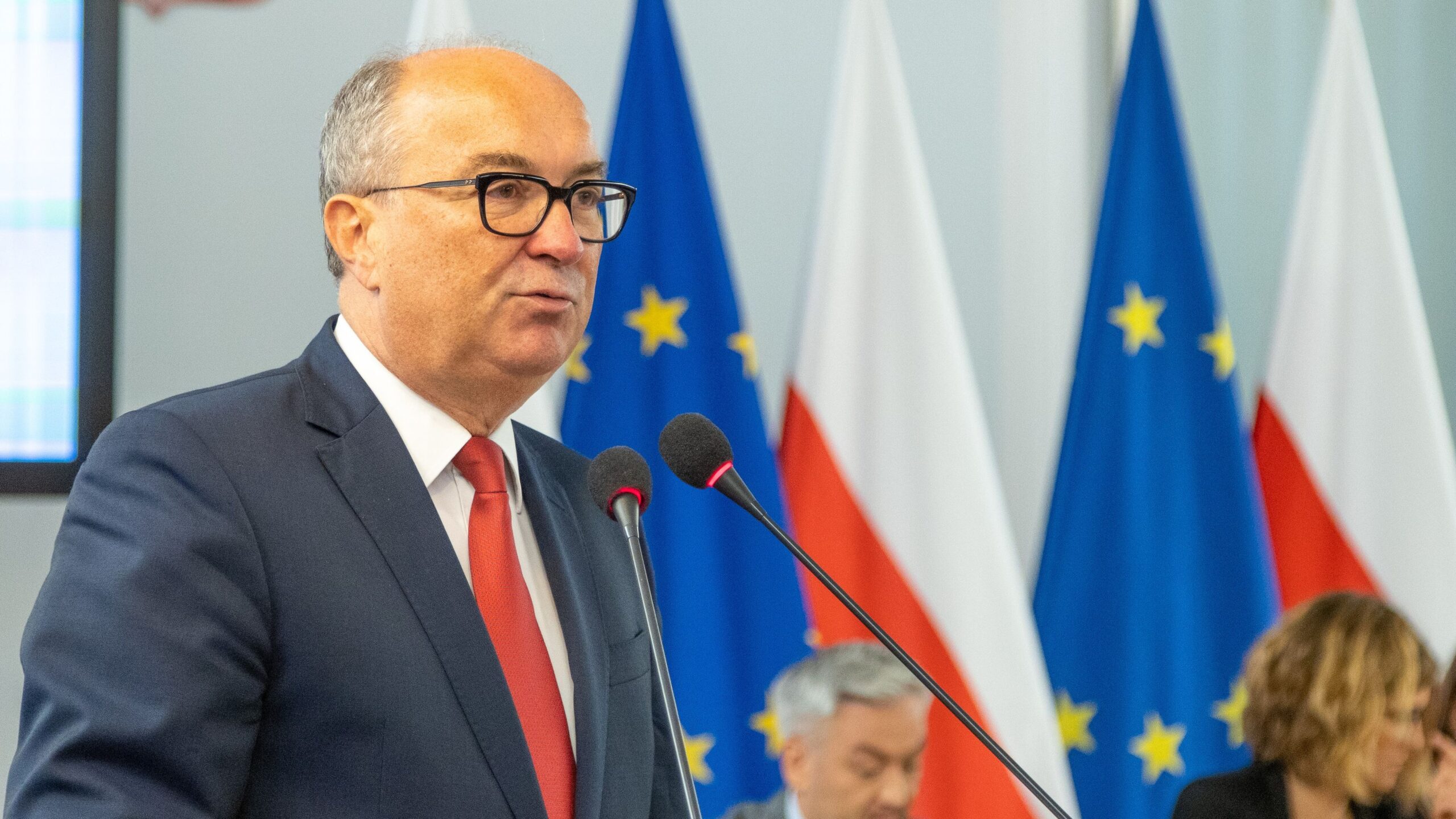 Na zdjęciu widać mężczyznę w okularach, który przemawia przy mikrofonie, a w tle znajdują się flagi Unii Europejskiej i Polski.