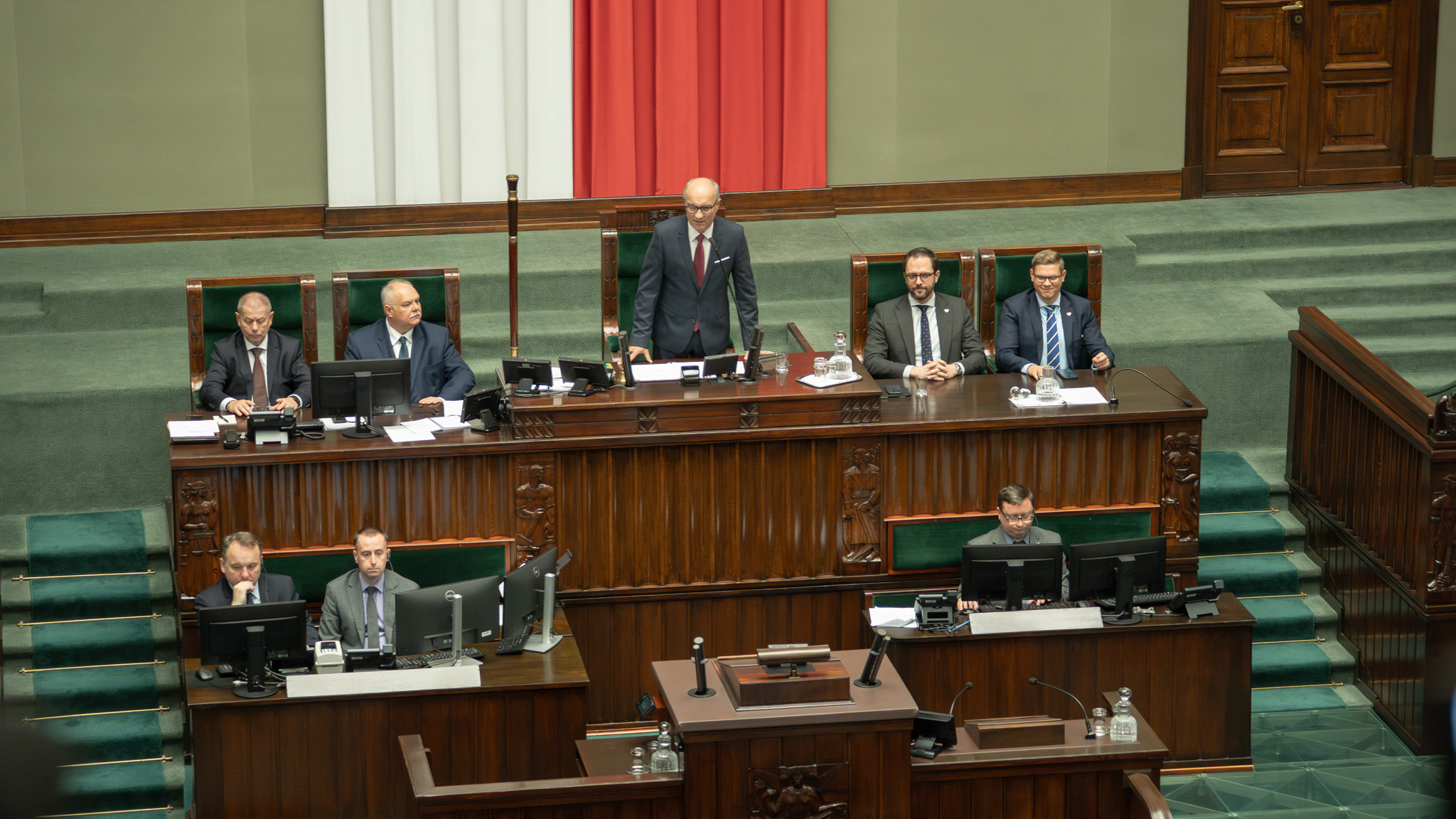 Mężczyzna w garniturze stoi przy mównicy na podium w sali plenarnej Sejmu, otoczony innymi urzędnikami siedzącymi przy biurkach.