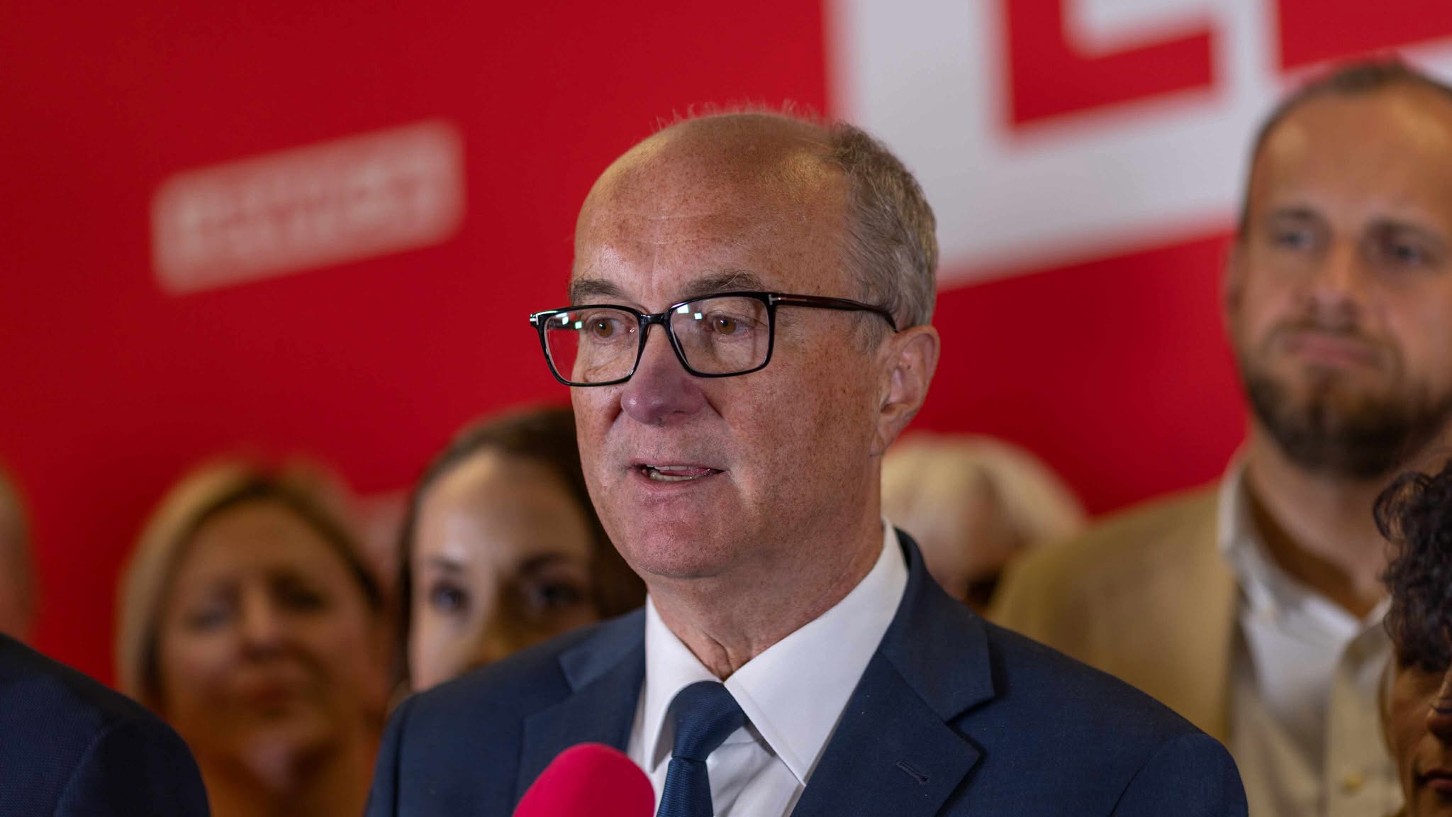 Mężczyzna w granatowym garniturze i czarnych okularach przemawia na Kongresie Nowej Lewicy, na czerwonym tle z logotypem partii.