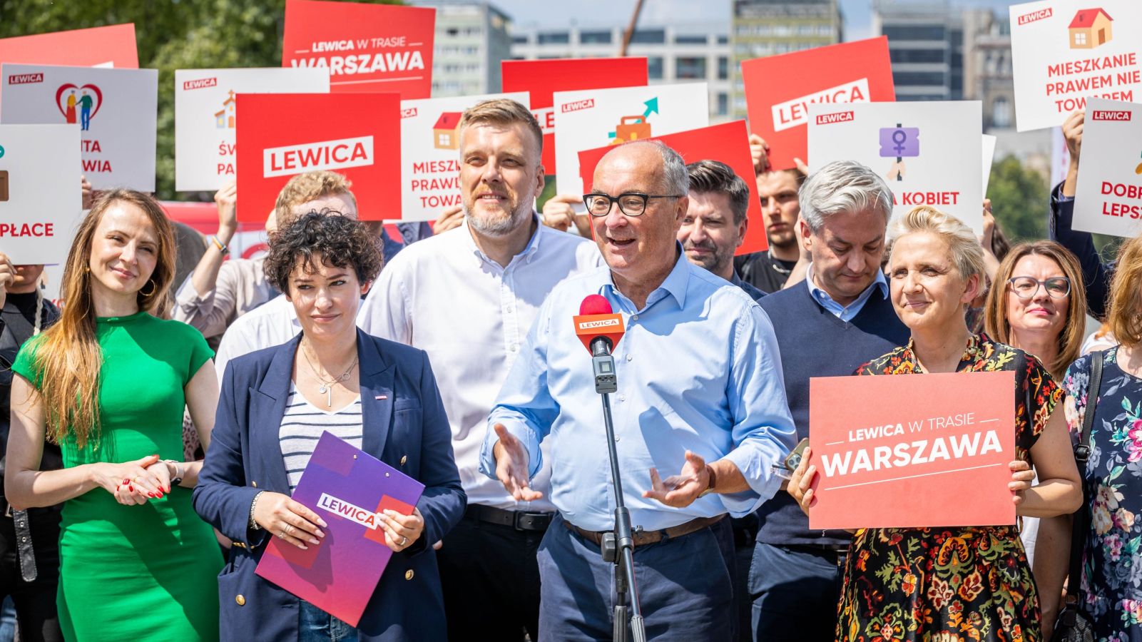 Grupa osób z plakatami Lewica na tle miasta, na pierwszym planie mężczyzna w błękitnej koszuli przemawia do mikrofonu podczas inauguracji Wakacyjnej Trasy Lewicy.