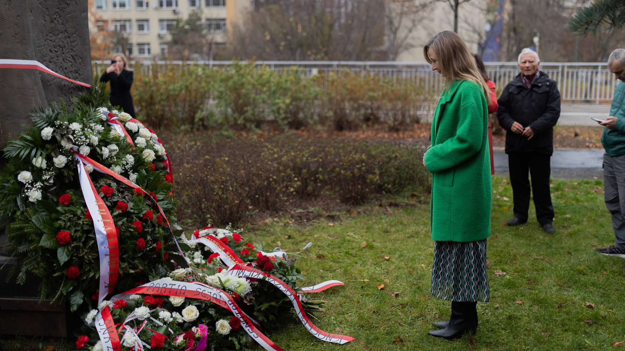 Kobieta w zielonym płaszczu stoi na trawie przed kwiatowymi wieńcami złożonymi w hołdzie podczas obchodów 106. rocznicy powołania rządu Ignacego Daszyńskiego.