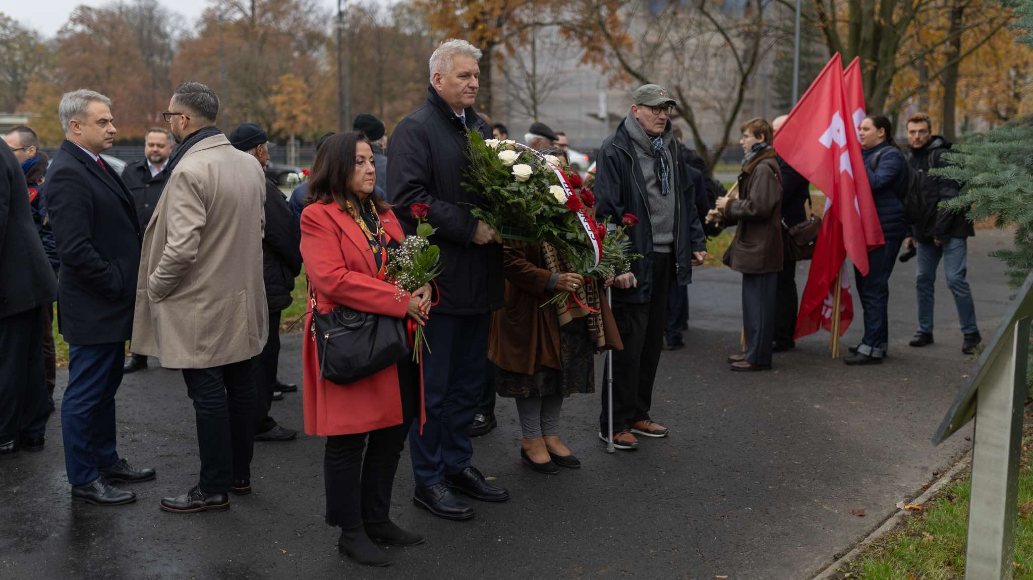 Grupa uczestników stoi na zewnątrz, składa kwiaty i trzyma czerwone flagi podczas obchodów 106. rocznicy powołania rządu Ignacego Daszyńskiego.