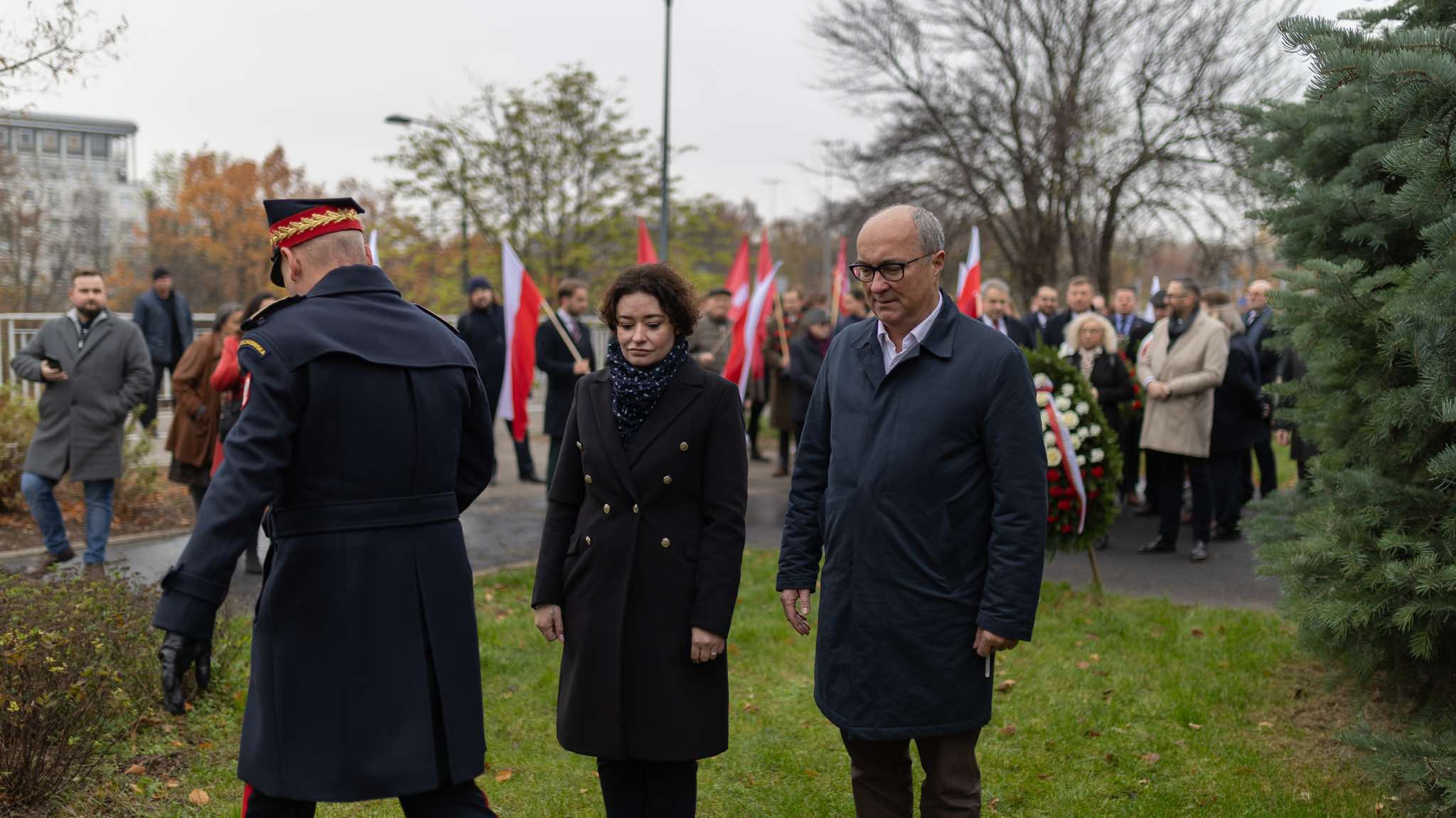 Uczestnicy uroczystości z okazji 106. rocznicy powołania rządu Ignacego Daszyńskiego idą z flagami; na pierwszym planie para — kobieta w czarnym płaszczu i mężczyzna w granatowym, po lewej stronie stojący w mundurze funkcjonariusz.