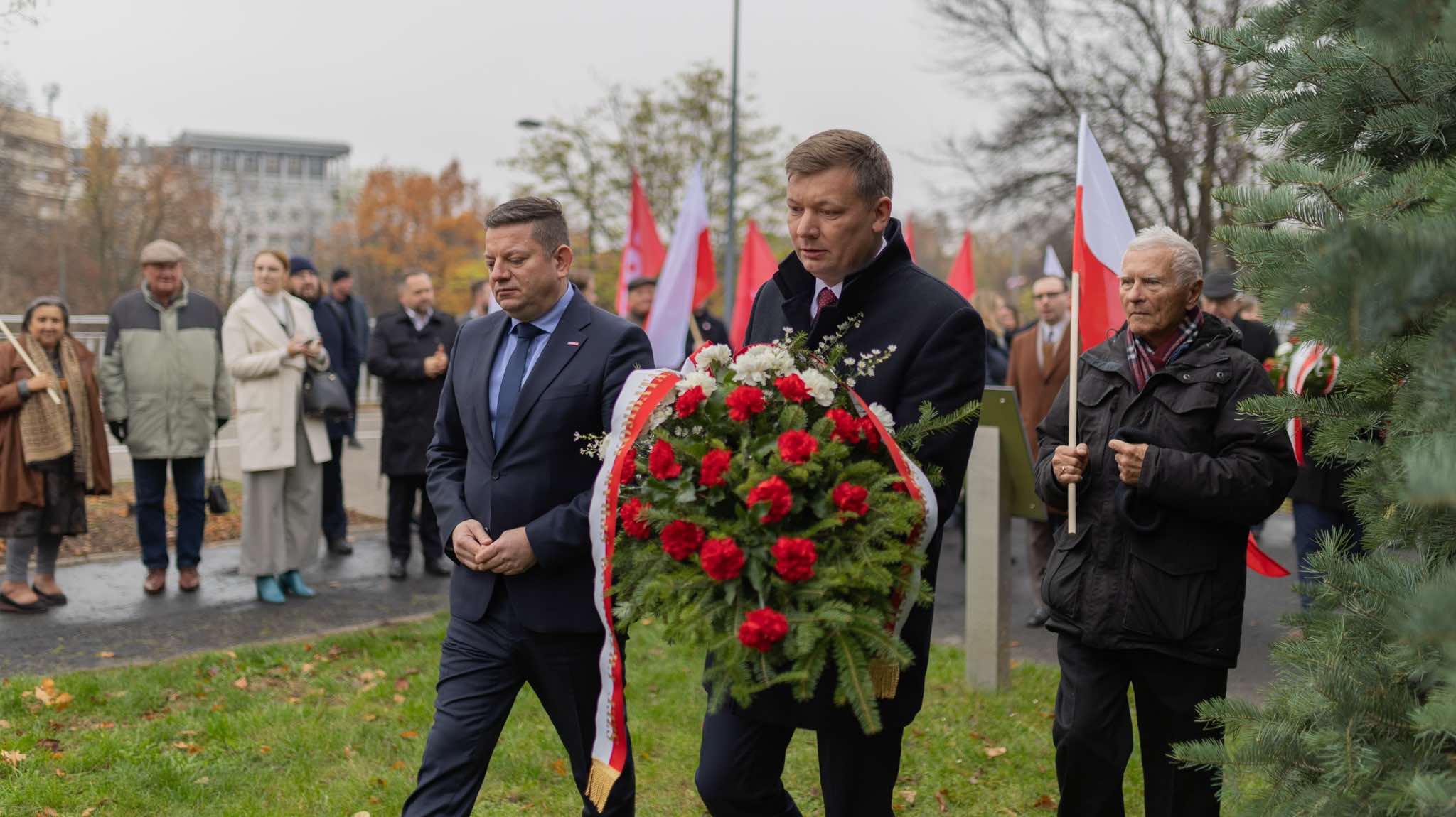 Dwóch mężczyzn w garniturach niesie okazały wieniec z czerwonych i białych kwiatów podczas uroczystości upamiętniającej 106. rocznicę powołania rządu Ignacego Daszyńskiego, na tle zgromadzonych uczestników i flag państwowych.