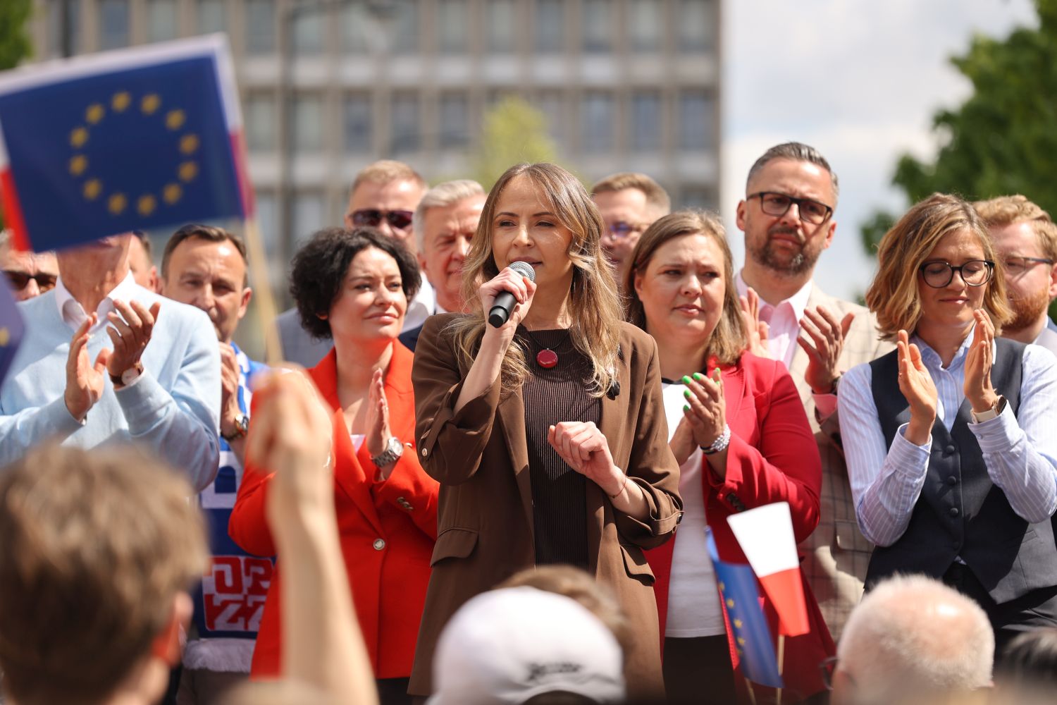 Kobieta przemawia do mikrofonu na plenerowym zgromadzeniu z flagami UE i Polski podczas Święta Pracy 2025.