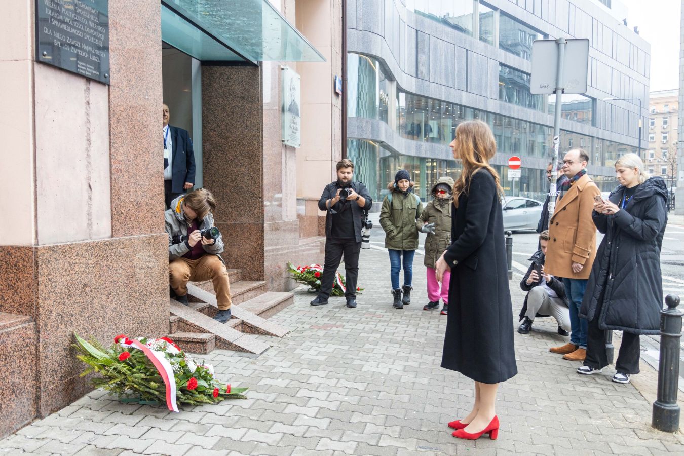 Kobieta w czarnym płaszu i czerwonych szpilkach stoi przed wejściem do budynku i przemawia do zgromadzonych dziennikarzy i przechodniów, podczas gdy na chodniku leżą kwietne wieńce.