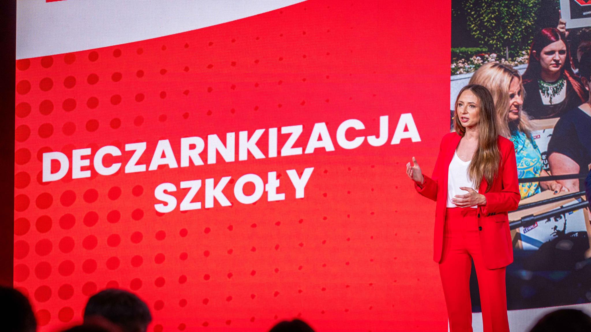 Kobieta w czerwonym garniturze przemawia na scenie przed dużym czerwonym ekranem z białymi napisami.