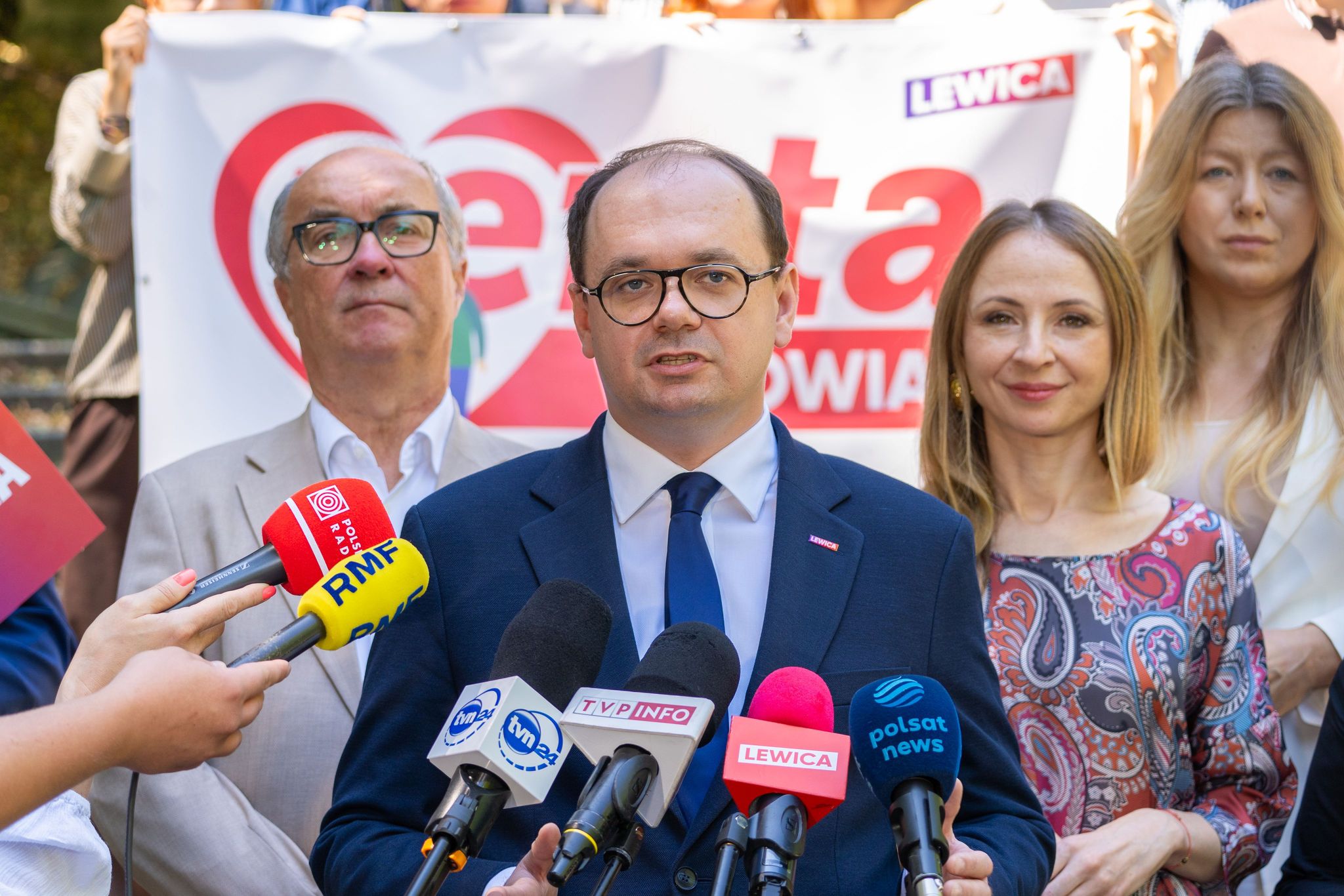Mężczyzna w niebieskim garniturze i okularach przemawia na konferencji prasowej, otoczony dziennikarzami i mikrofonami, a w tle widać baner Lewicy.