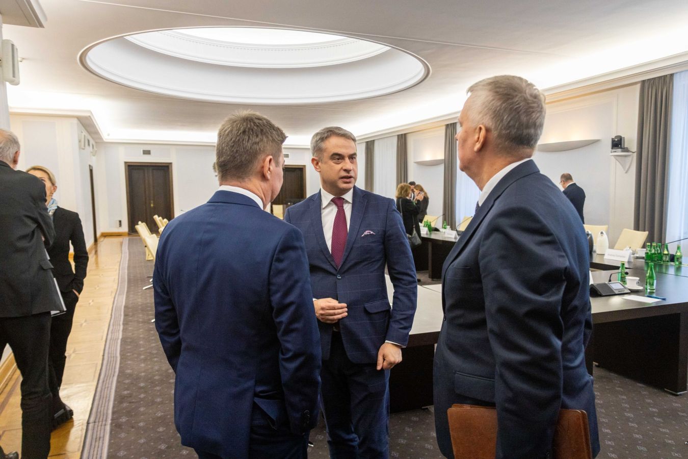 Trzech mężczyzn w garniturach rozmawia ze sobą w sali konferencyjnej, stojąc obok długiego stołu z dokumentami i zielonymi butelkami wody.