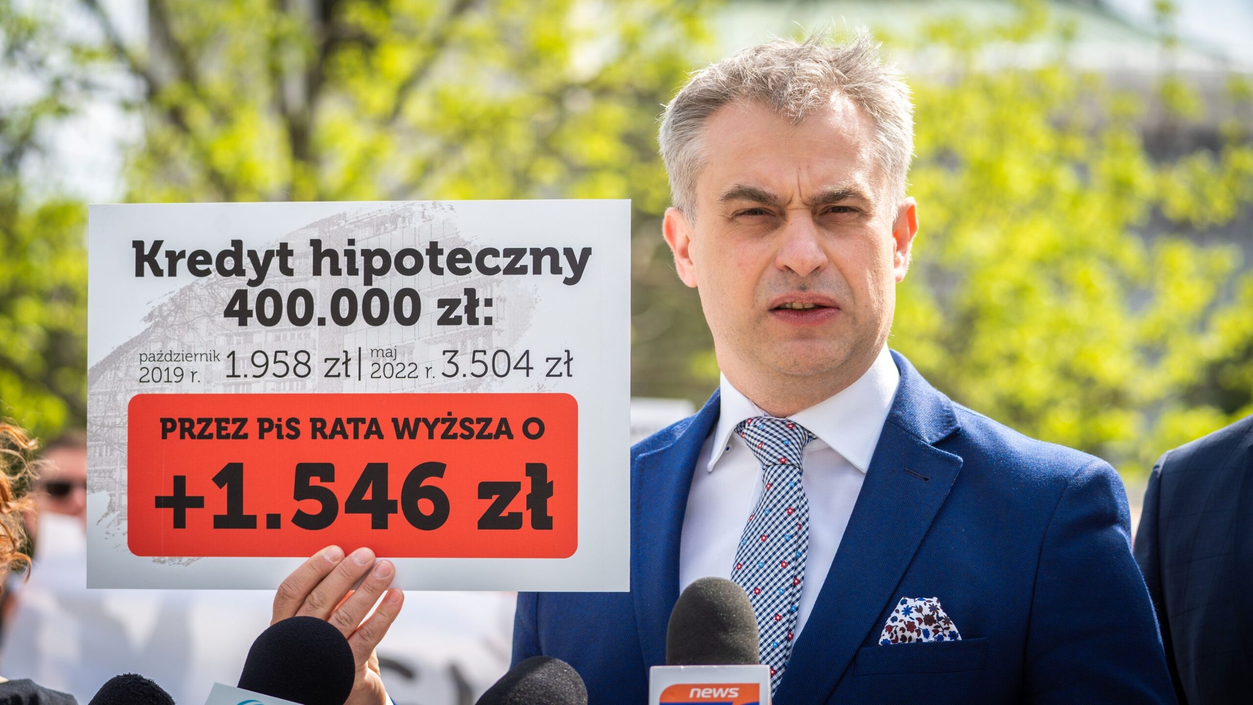 Mężczyzna w garniturze trzyma plakat dotyczący wzrostu rat kredytu hipotecznego, stojąc w otoczeniu zieleni.
