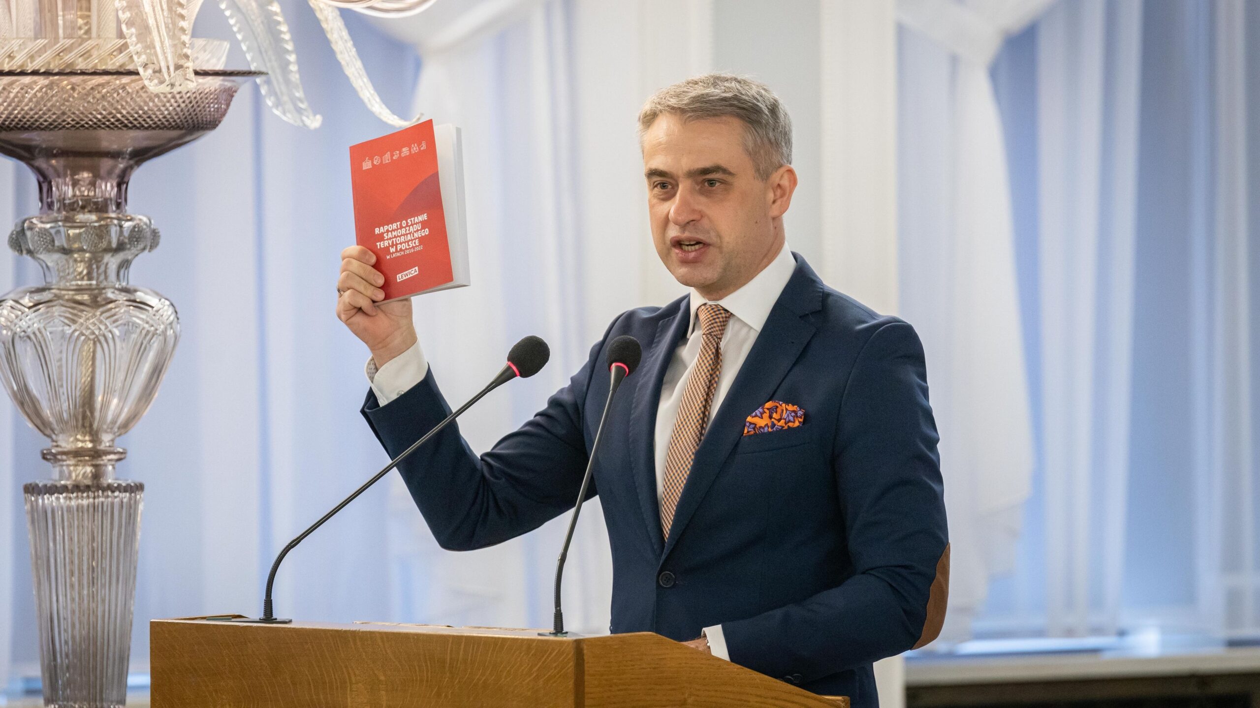Mężczyzna w granatowym garniturze z pomarańczową poszetką stoi za mównicą, trzymając w lewej ręce czerwono‑biały podręcznik, a przed nim są mikrofony; w tle widoczny jest duży kryształowy wazon.