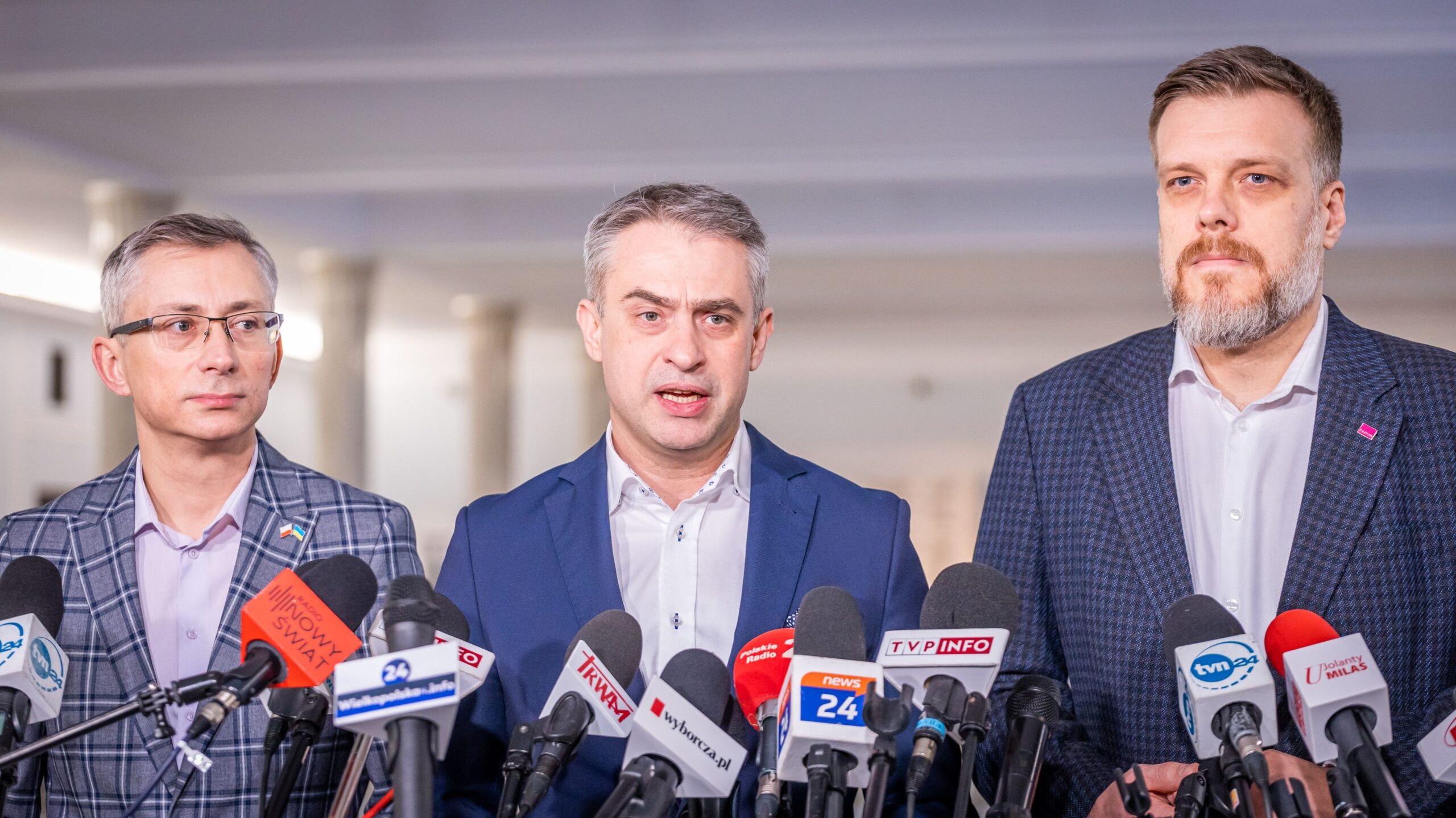 Na zdjęciu trzech mężczyzn stoi przed mikrofonami, prowadząc konferencję prasową.