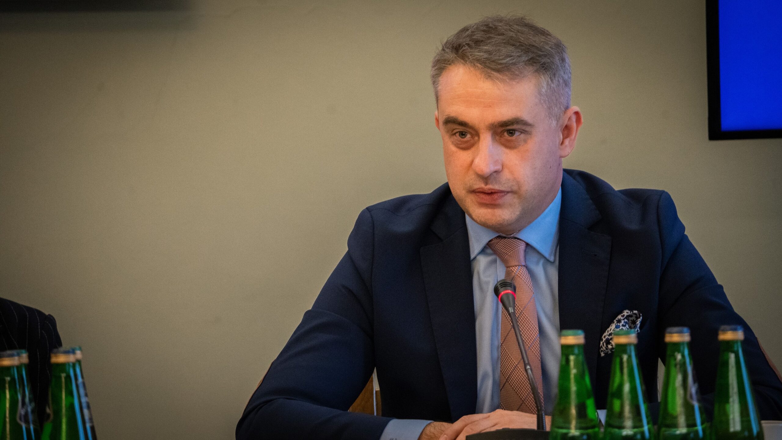 Mężczyzna w garniturze siedzi przy stole w trakcie spotkania, z mikrofonem przed sobą i butelkami wody w tle.