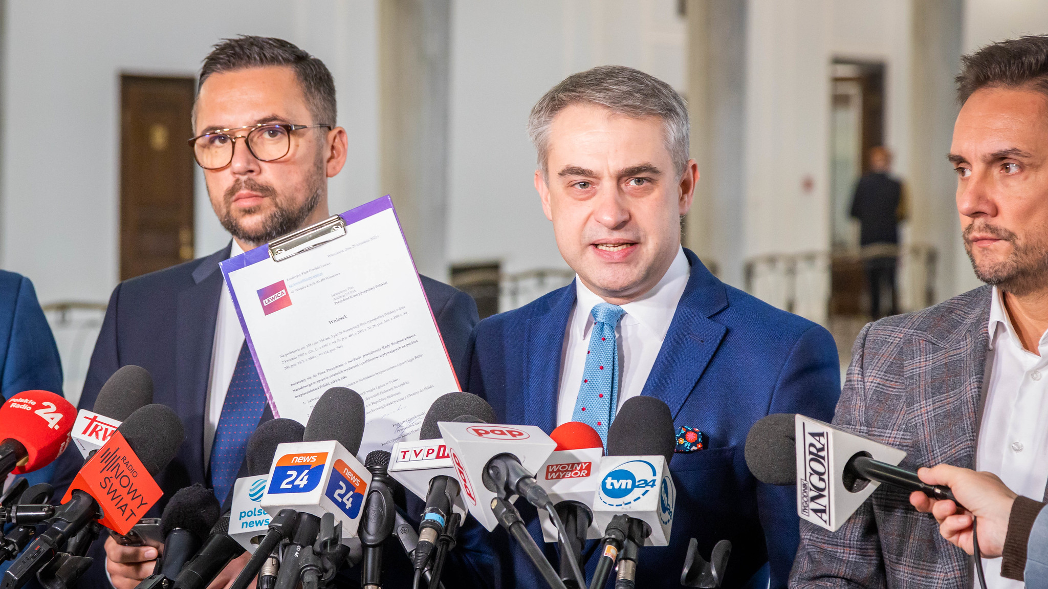 Na zdjęciu widoczna jest konferencja prasowa, na której mężczyzna w niebieskim garniturze trzyma dokument, a obok niego stoją inni mężczyźni, wokół są mikrofony mediów.