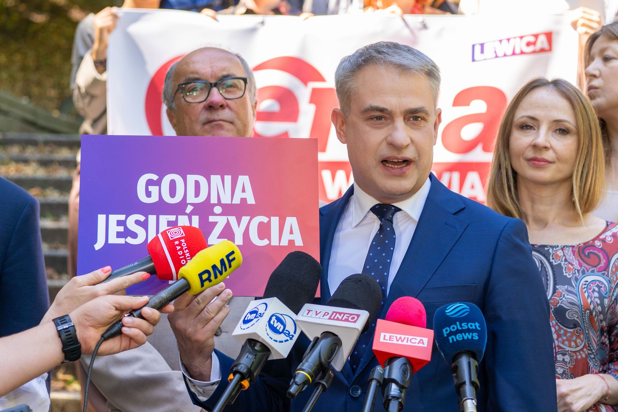 Mężczyzna w granatowym garniturze przemawia do mediów podczas konferencji prasowej, otoczony mikrofonami, z widocznym plakatem „GODNA JESIEŃ ŻYCIA” w tle.