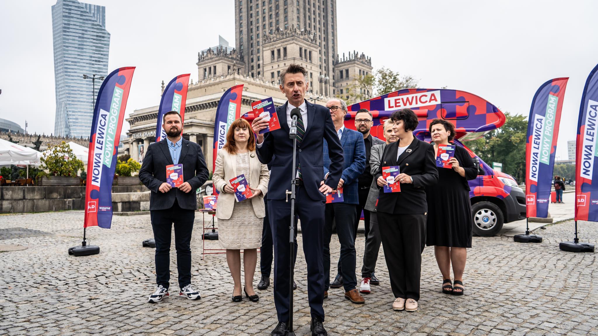 Mężczyzna w garniturze przemawia z mikrofonem na plenerowej konferencji Nowej Lewicy, otoczony działaczami trzymającymi ulotki i banery z napisem Nowa Lewica oraz kampanijny samochód w tle.