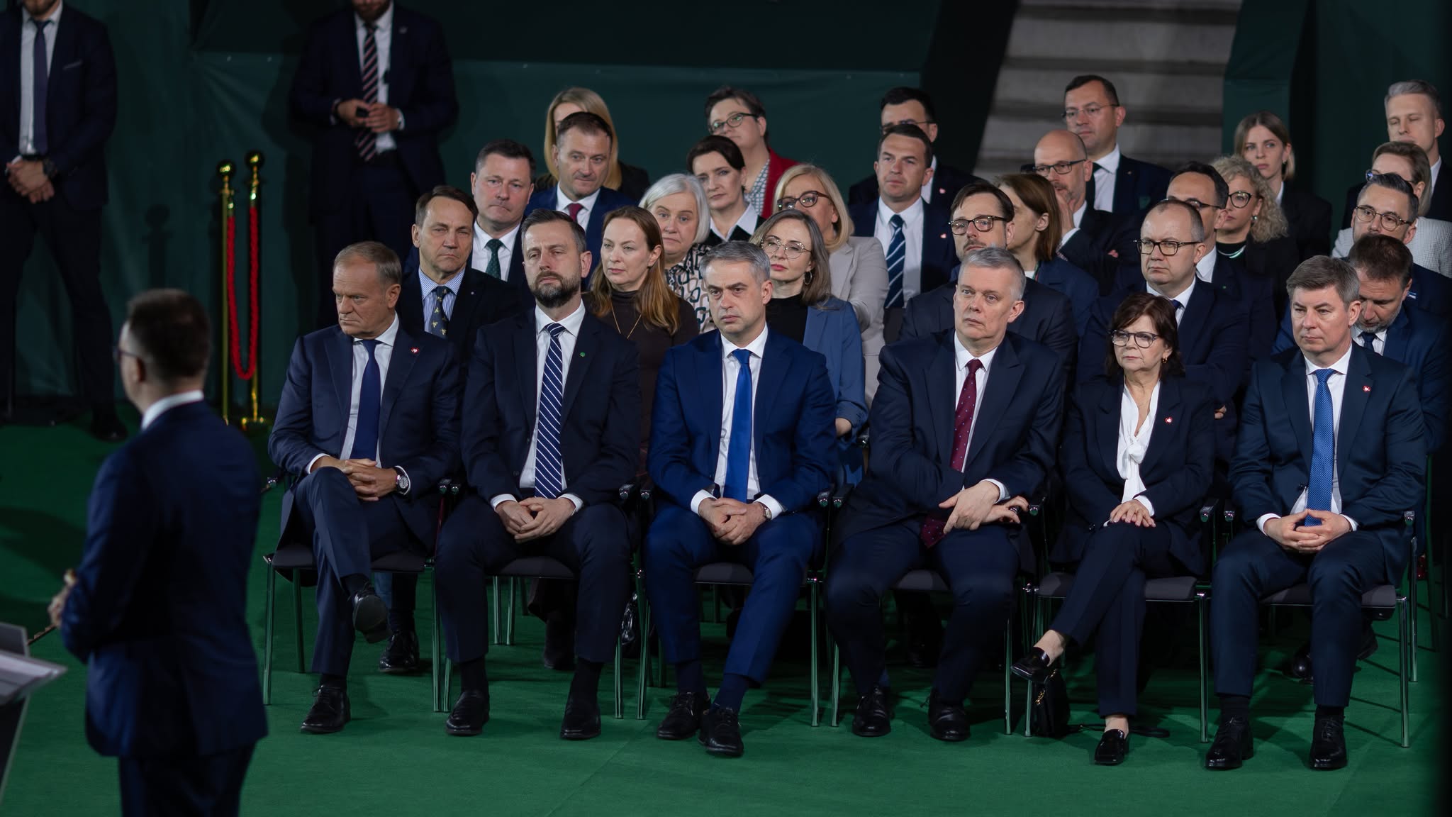 Grupa polityków i gości w garniturach siedzi na zielonej widowni podczas uroczystej ceremonii upamiętniającej 1000. rocznicę koronacji Bolesława Chrobrego.