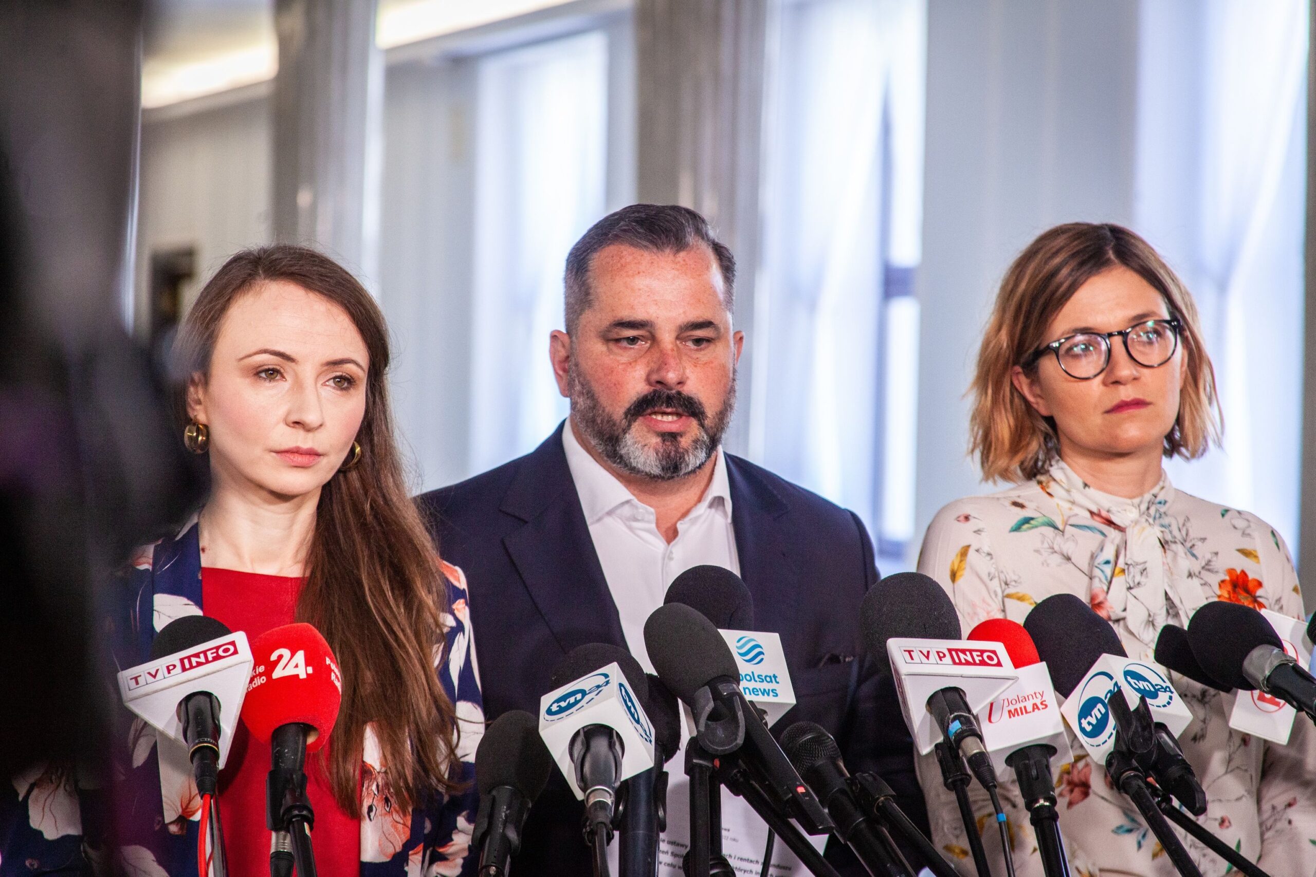 Trzy osoby stoją przed mikrofonami podczas konferencji prasowej w jasnym pomieszczeniu.