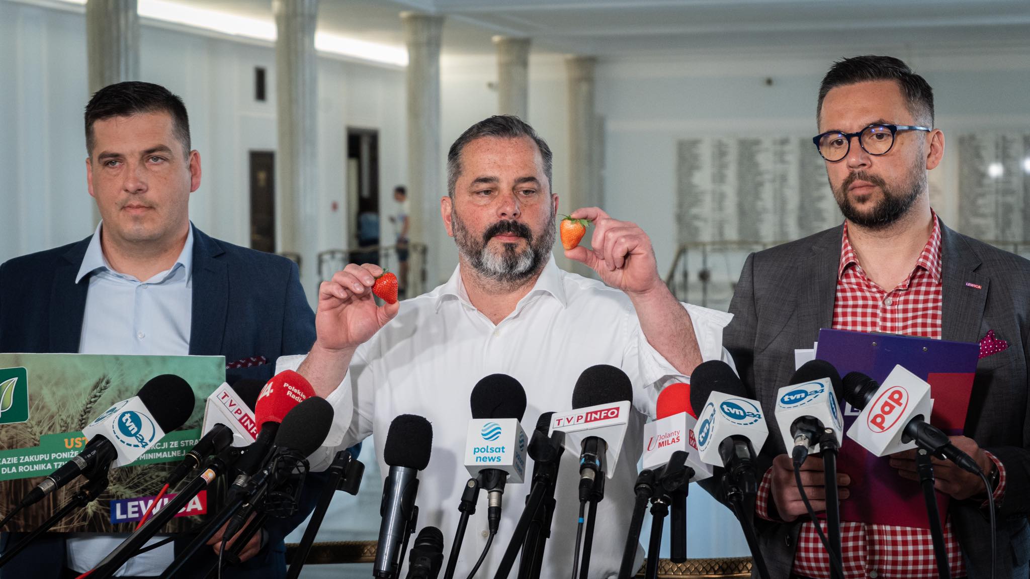 Trzech mężczyzn w garniturach podczas konferencji prasowej, środkowy pokazuje dwie truskawki, a przed nimi liczne mikrofony z logotypami mediów.