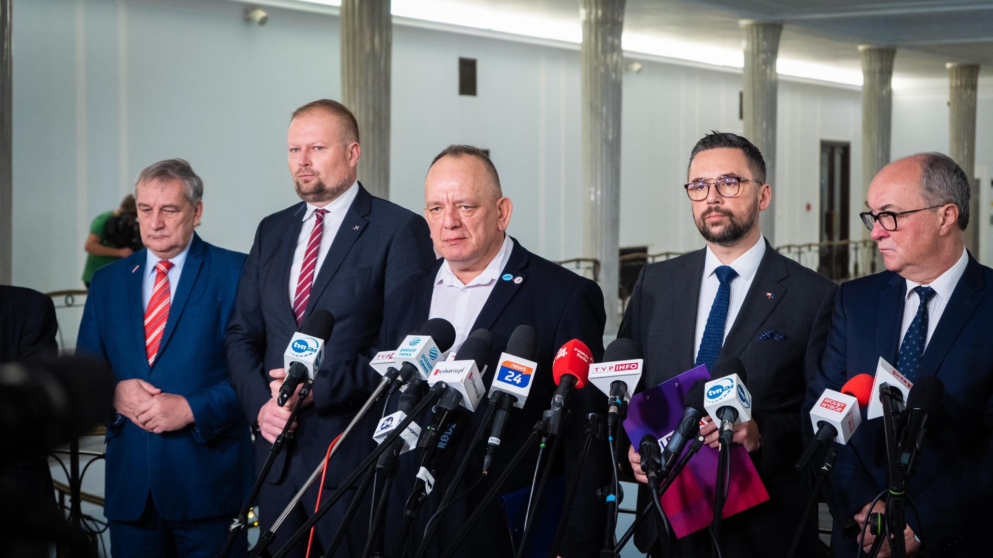 Na zdjęciu znajduje się grupka mężczyzn w garniturach, stojących przed mikrofonami podczas konferencji prasowej w budynku publicznym.