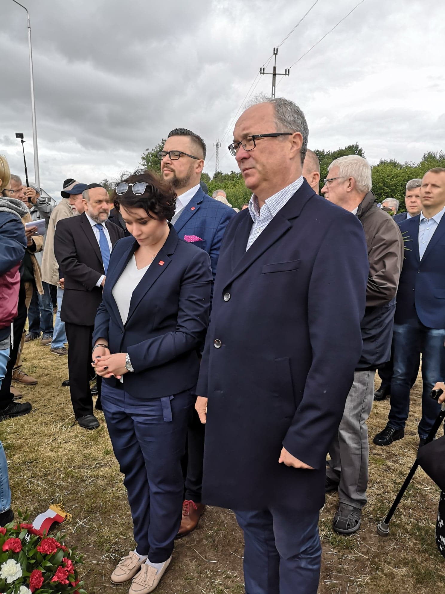 Grupa osób w garniturach i marynarkach stoi na otwartej przestrzeni podczas uroczystości upamiętniającej zamordowanych Żydów w Jedwabnem, przed złożonymi kwiatami.