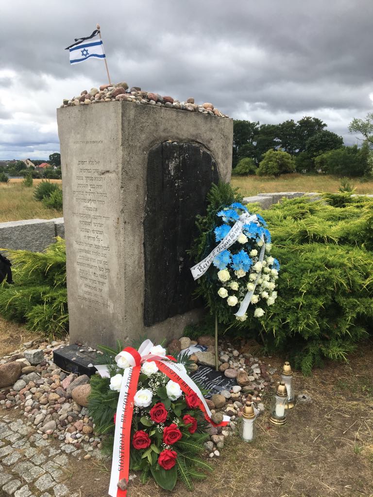 Monument upamiętniający zamordowanych Żydów w Jedwabnem: kamienny obelisk z tablicą, nad którym unosi się flaga Izraela, otoczony wieńcami, kwiatami i zapalonymi zniczami.