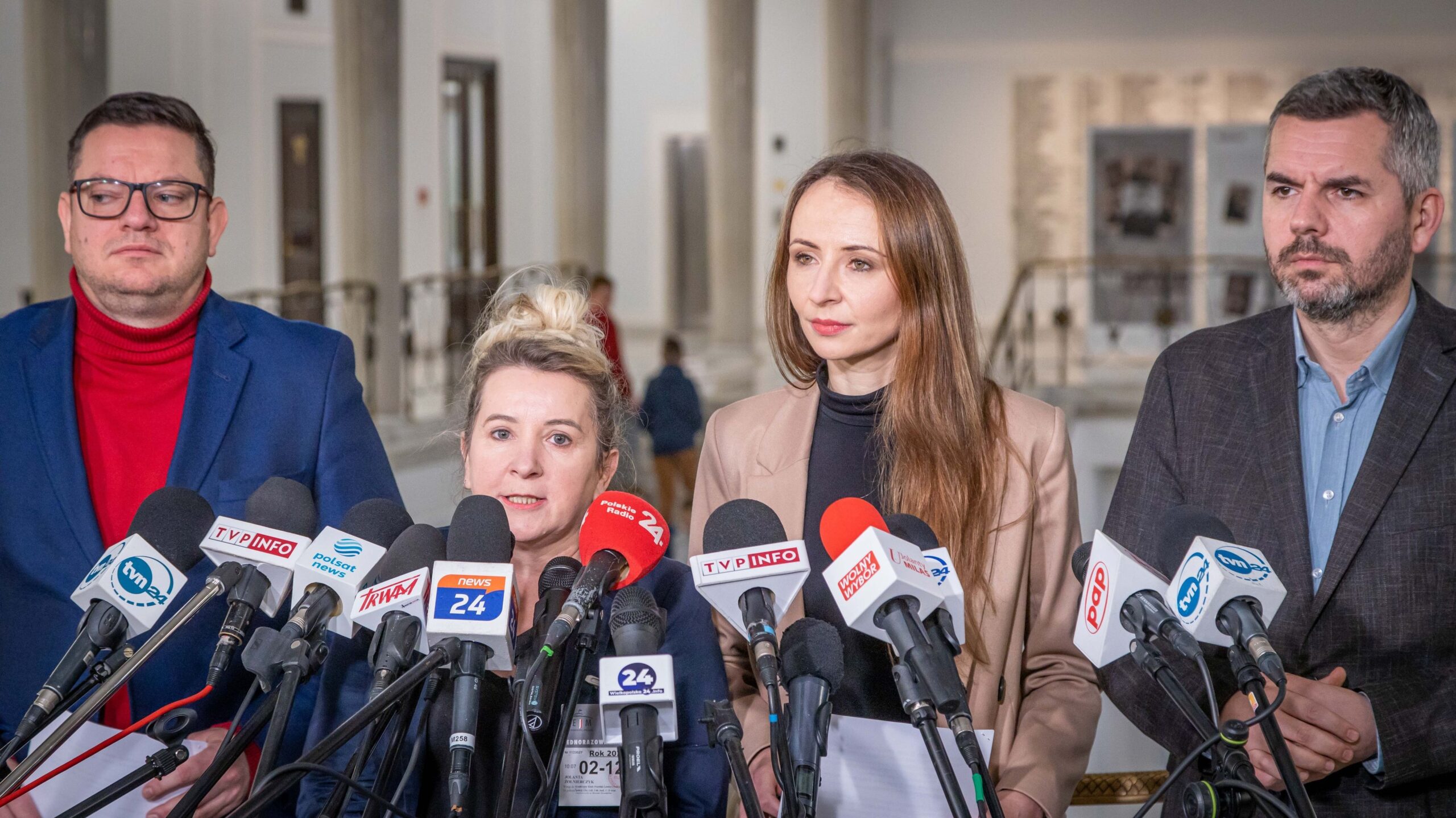 Zdjęcie przedstawia czterech polityków podczas konferencji prasowej, stojących przy mikrofonach w tle nowoczesnego wnętrza.
