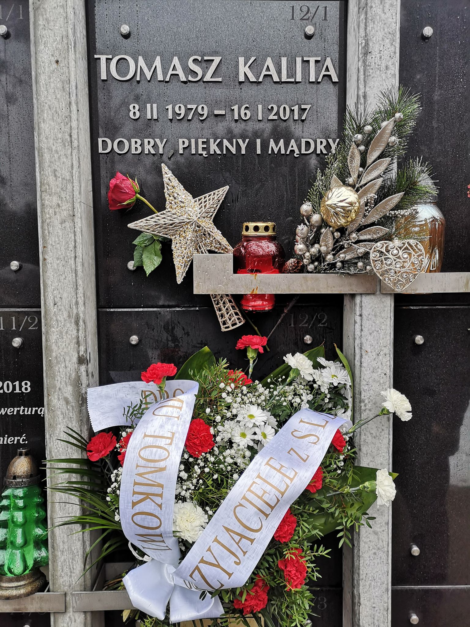 Tablica pamięci na grobie z napisem i datami, udekorowana kwiatami i innymi ozdobami.