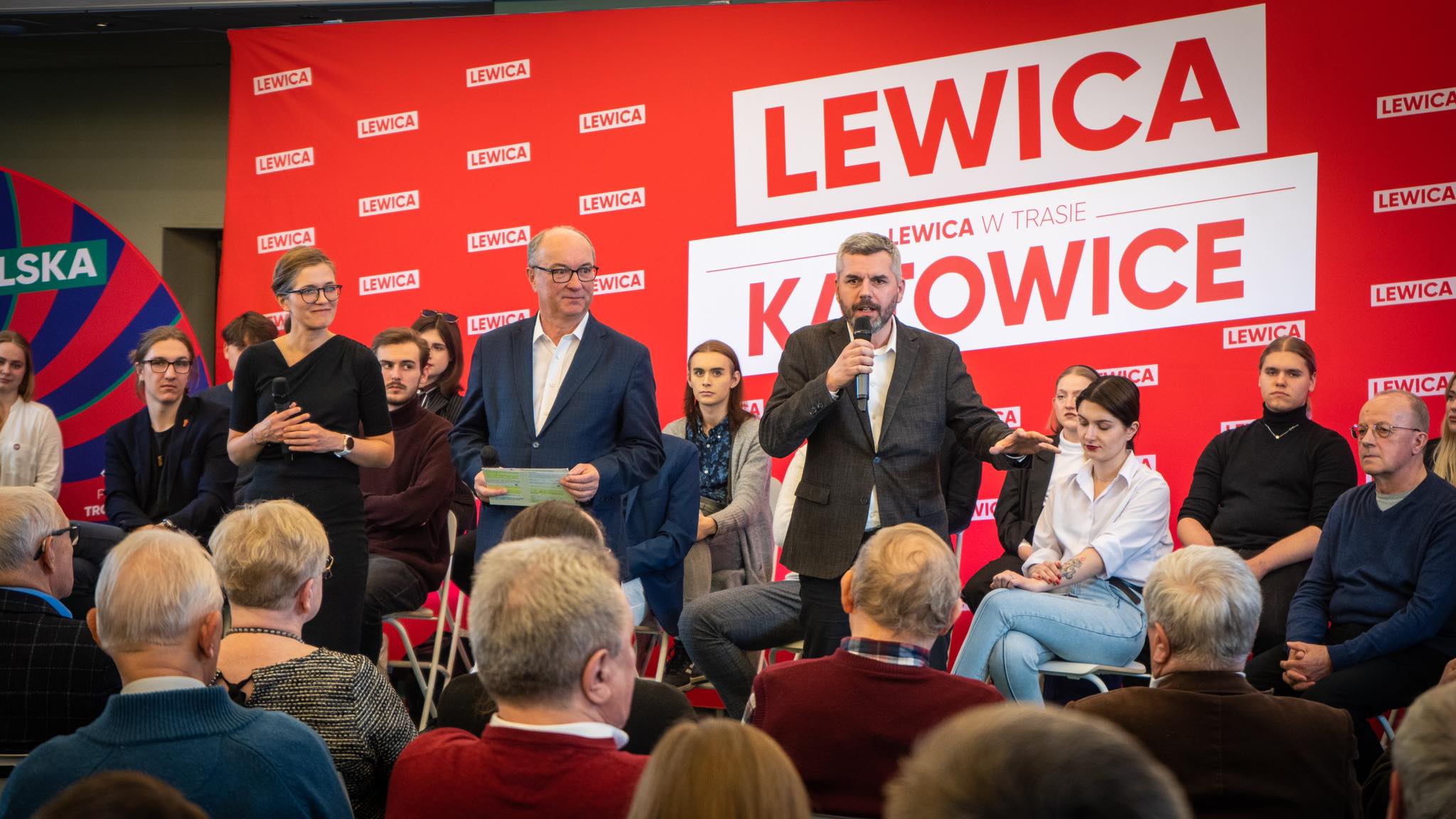 Panel Lewicy w Katowicach: mężczyzna z mikrofonem przemawia na scenie w towarzystwie innych uczestników, na tle czerwonego baneru z napisem LEWICA i KATOWICE.