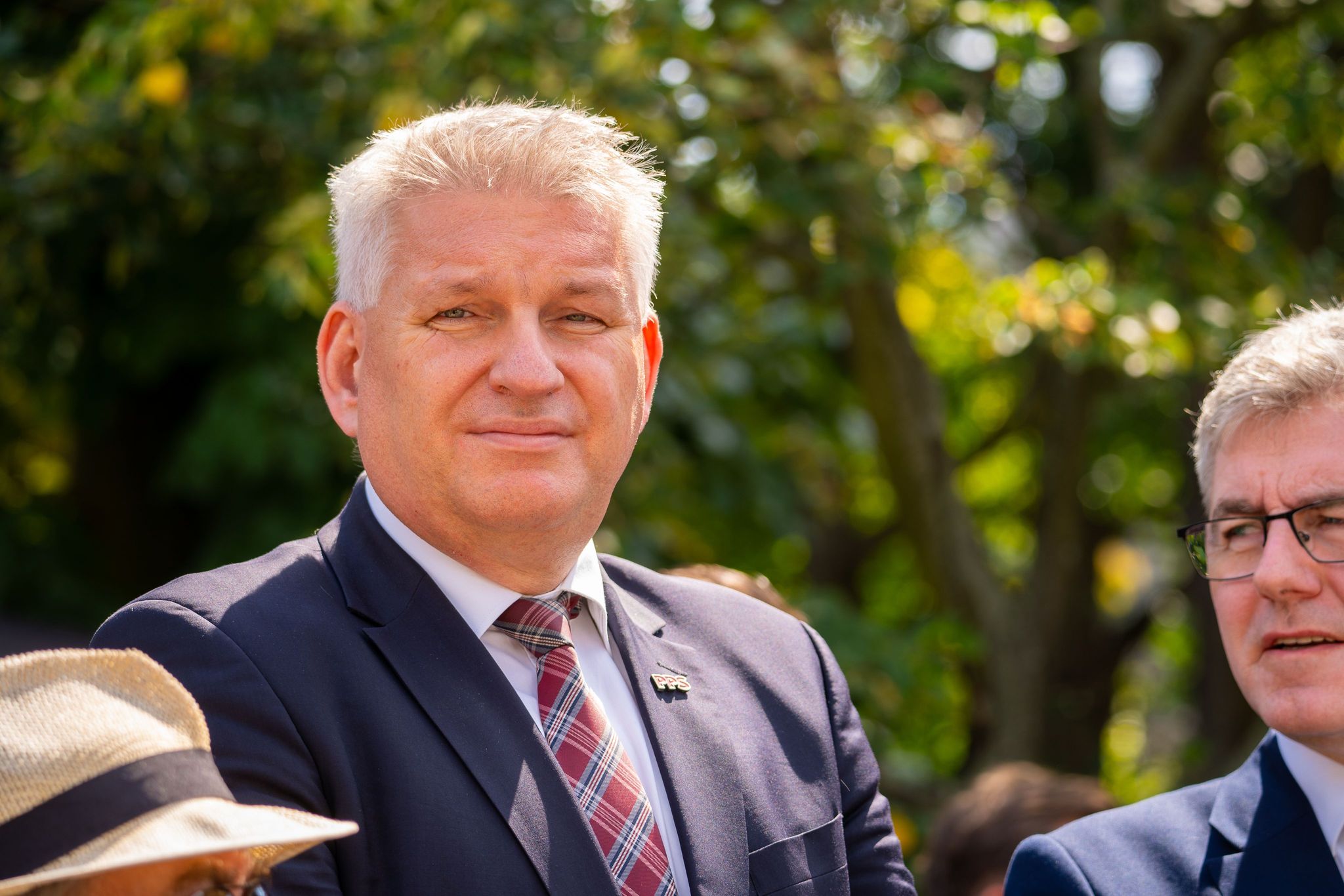 Mężczyzna w garniturze i kraciastym krawacie stoi na zewnątrz podczas obchodów 80. rocznicy Powstania Warszawskiego, w tle zielone drzewa.