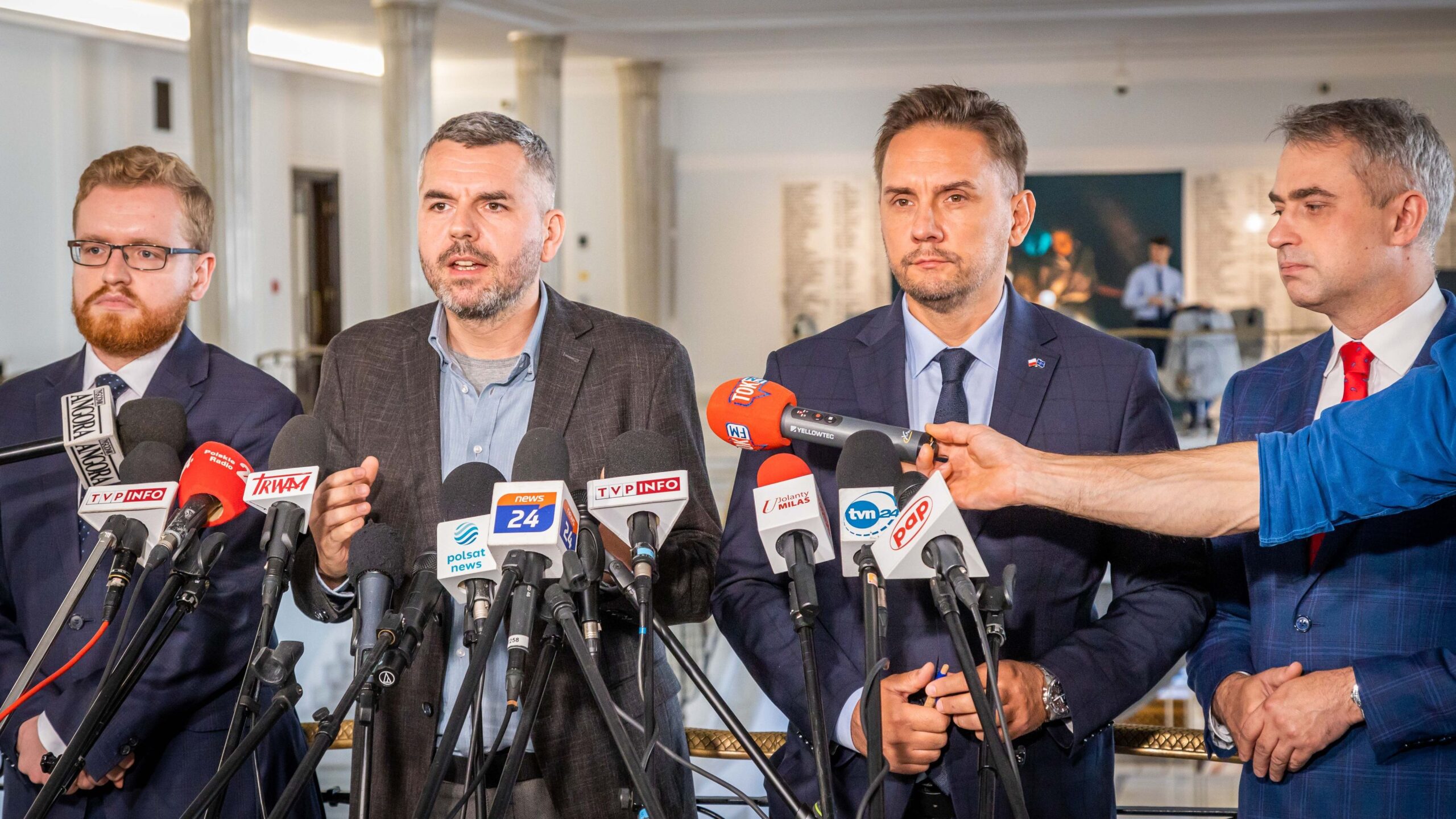 Na zdjęciu czterech mężczyzn w garniturach stoi przy mikrofonach na konferencji prasowej w urzędzie.