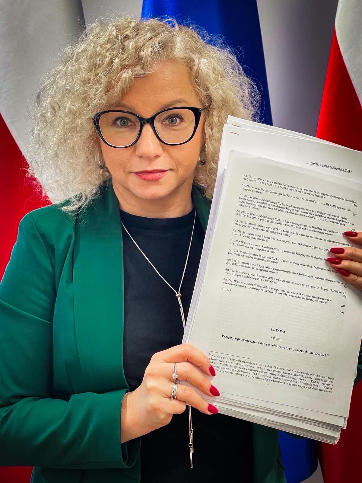 Kobieta z kręconymi jasnymi włosami i czarnymi okularami trzyma stos stron z projektem ustawy o rejestrowanych związkach partnerskich, stojąc na tle flag.