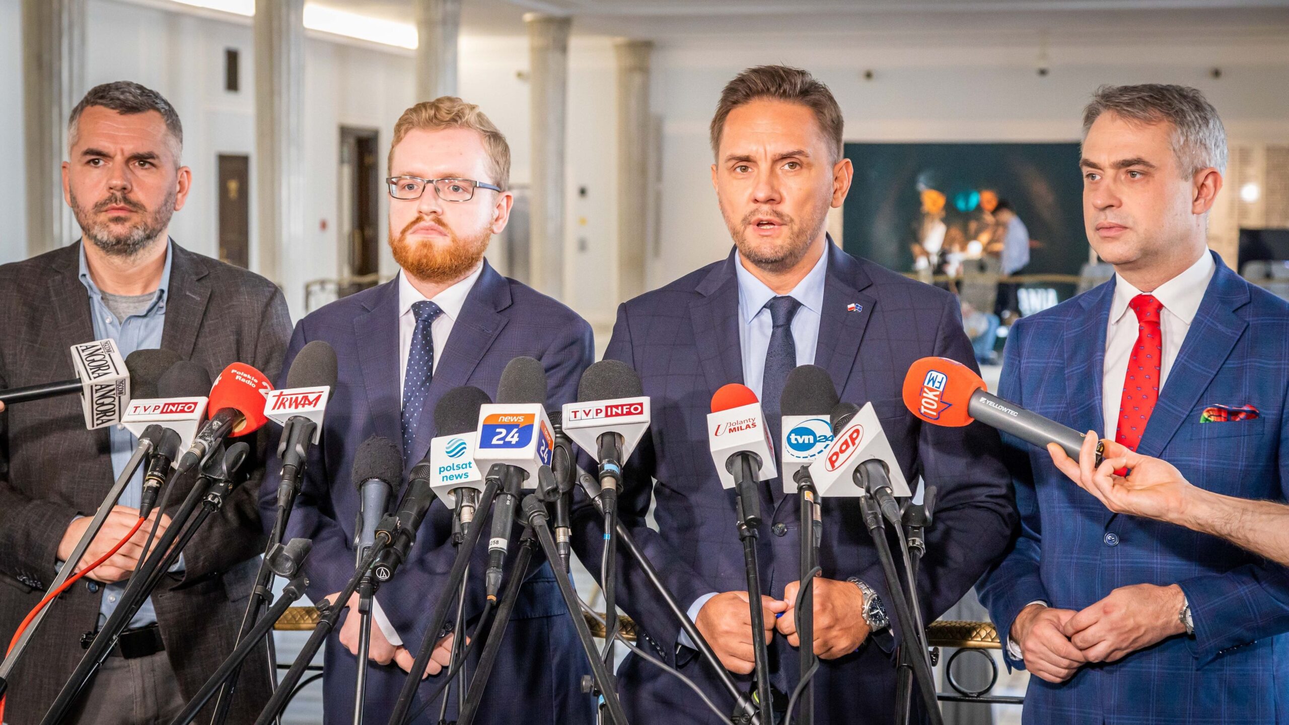 Czterech mężczyzn w garniturach stoi za mikrofonami podczas konferencji prasowej w eleganckim wnętrzu.