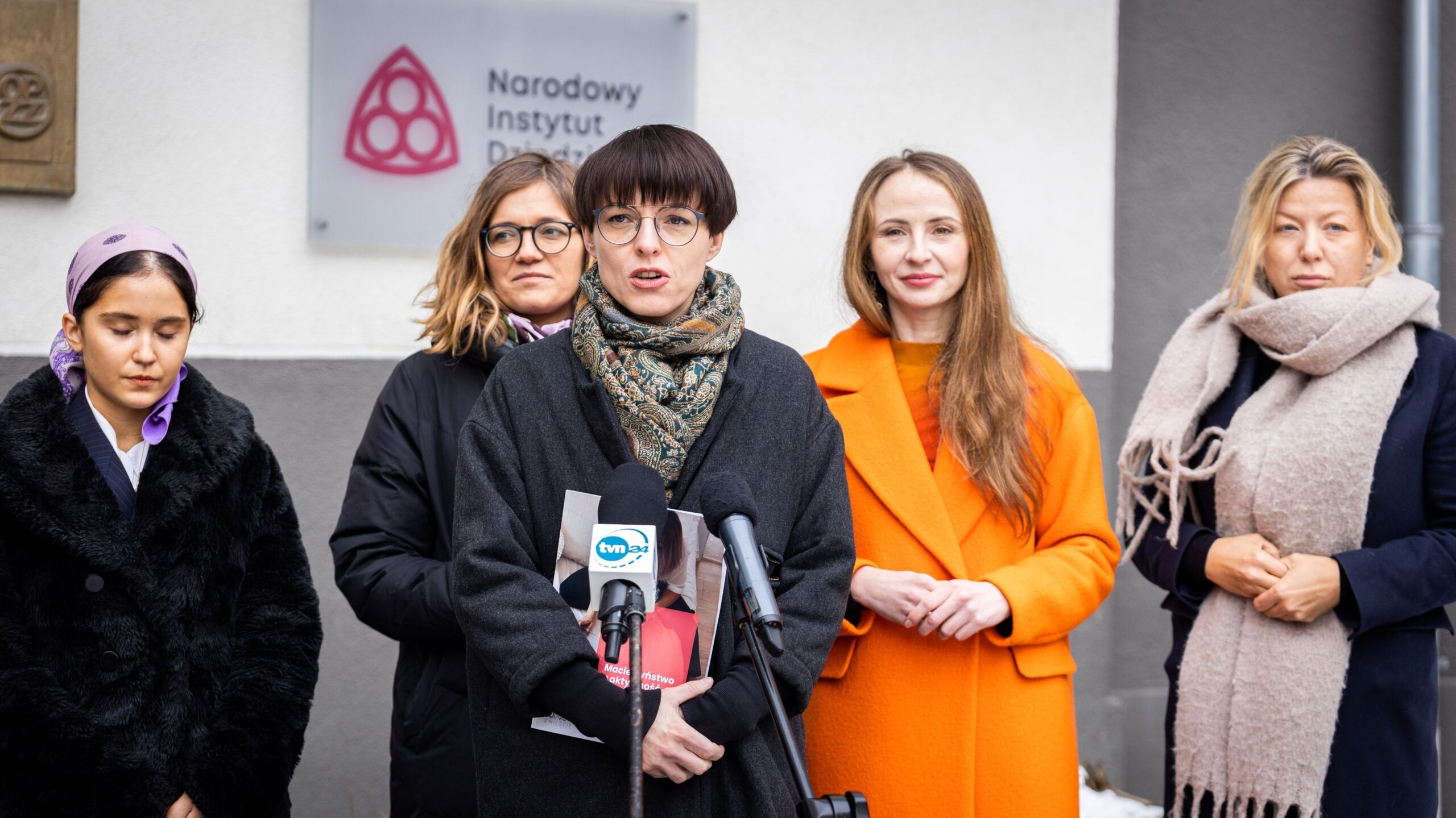 Na zdjęciu widoczne są pięć kobiet w różnych strojach stojących przed siedzibą Narodowego Instytutu Dziedzictwa, podczas konferencji prasowej.