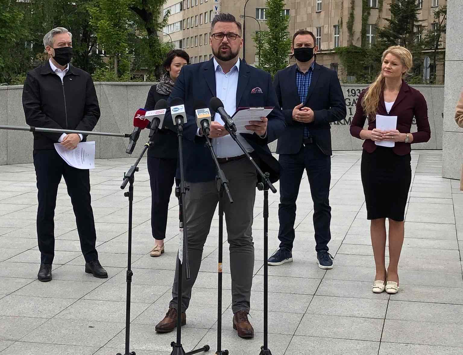 Mężczyzna w granatowym garniturze przemawia na plenerowej konferencji prasowej na placu, stojąc przed kilkoma mikrofonami, w towarzystwie współpracowników.