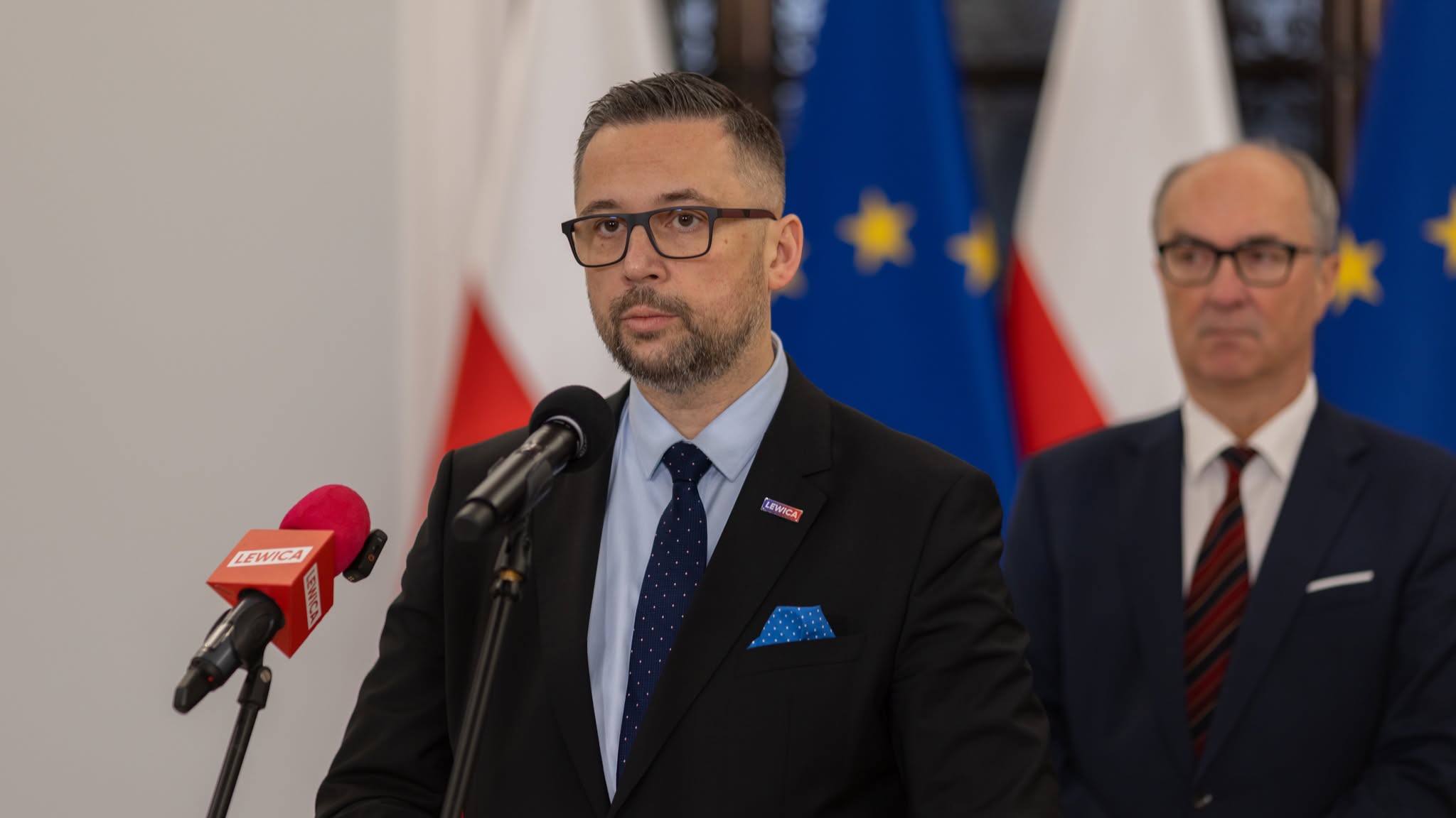 Polityk Lewicy w czarnym garniturze i okularach przemawia na konferencji prasowej, stojąc obok mikrofonów z logiem Lewicy, z flagami Polski i Unii w tle.