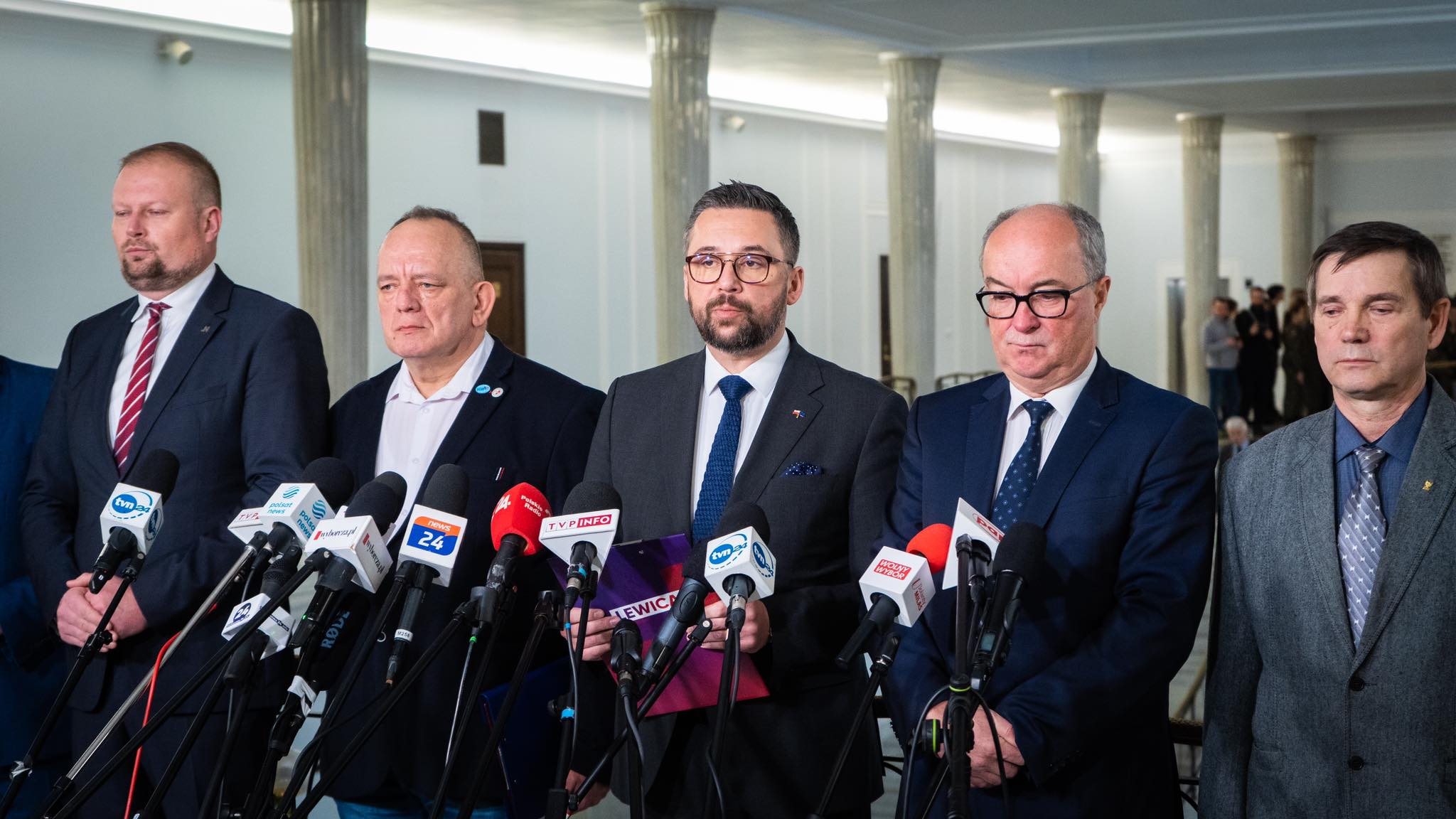 Grupa mężczyzn w garniturach stoi przy mównicy otoczonej mikrofonami, podczas konferencji prasowej.