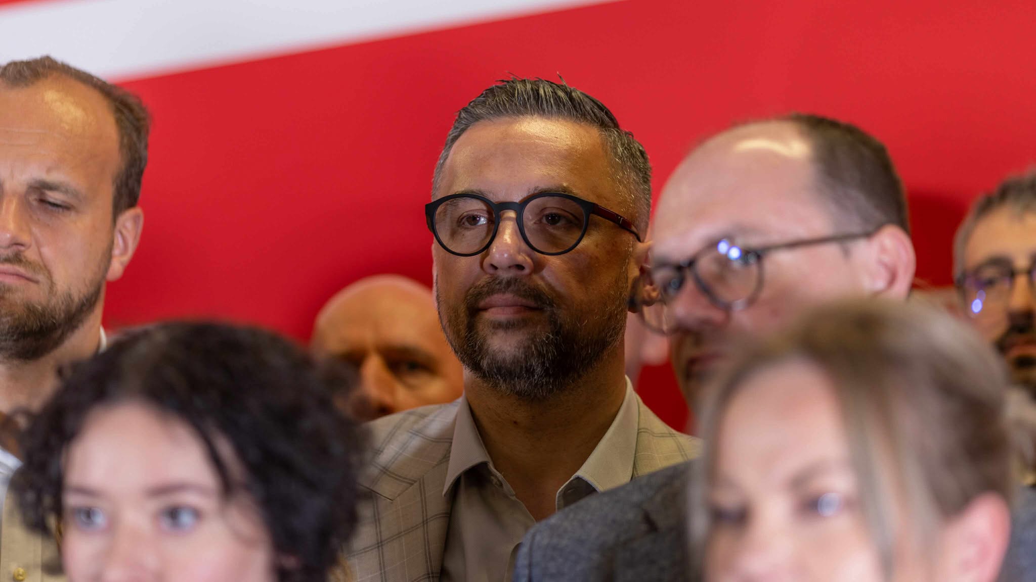 Mężczyzna z okrągłymi czarnymi okularami i krótką brodą stoi w tłumie podczas Kongresu Nowej Lewicy, na tle czerwonej sceny.