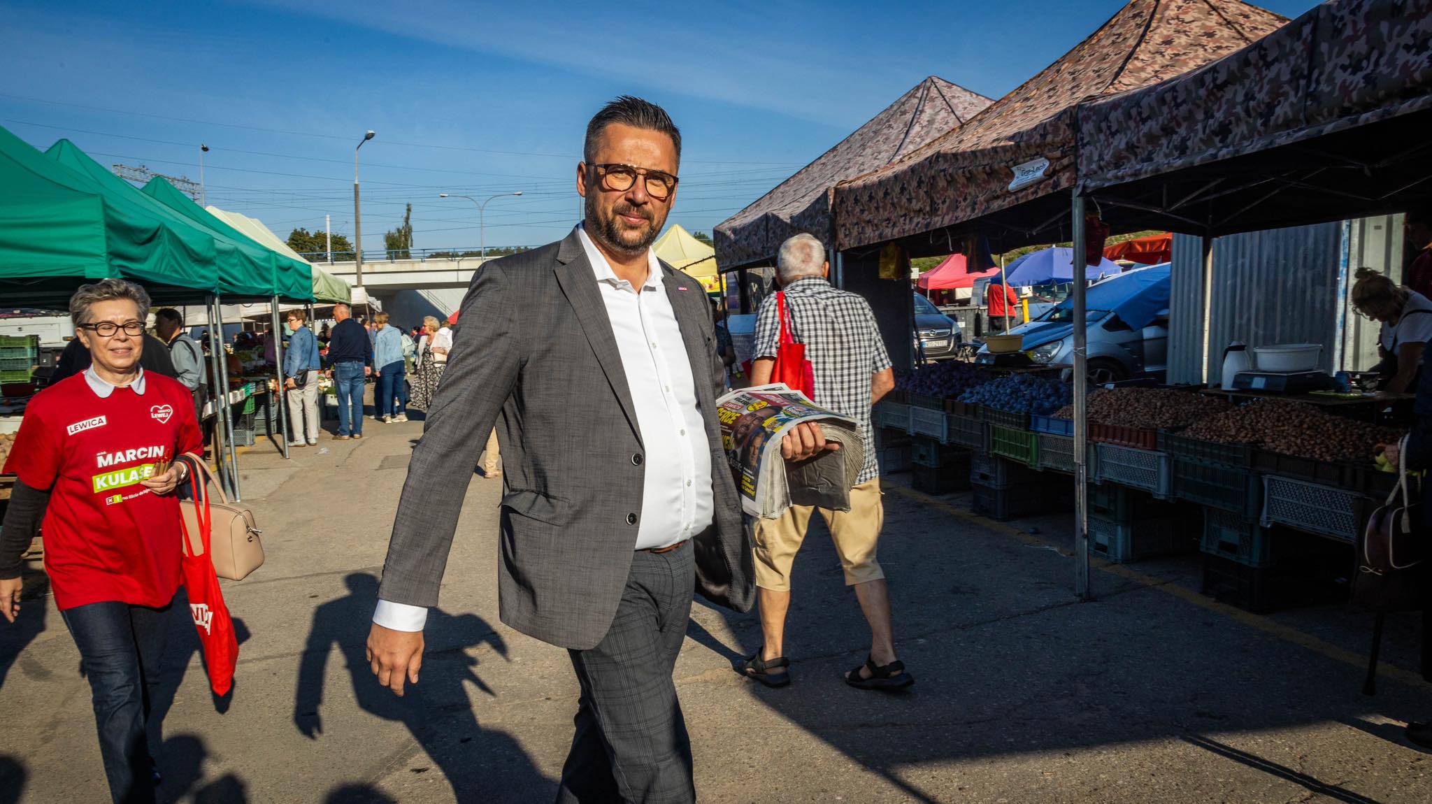 Mężczyzna w szarym garniturze przechodzi przez targ pod namiotami, trzyma gazetę, a w tle stoją stoiska z warzywami i ludzie robią zakupy.