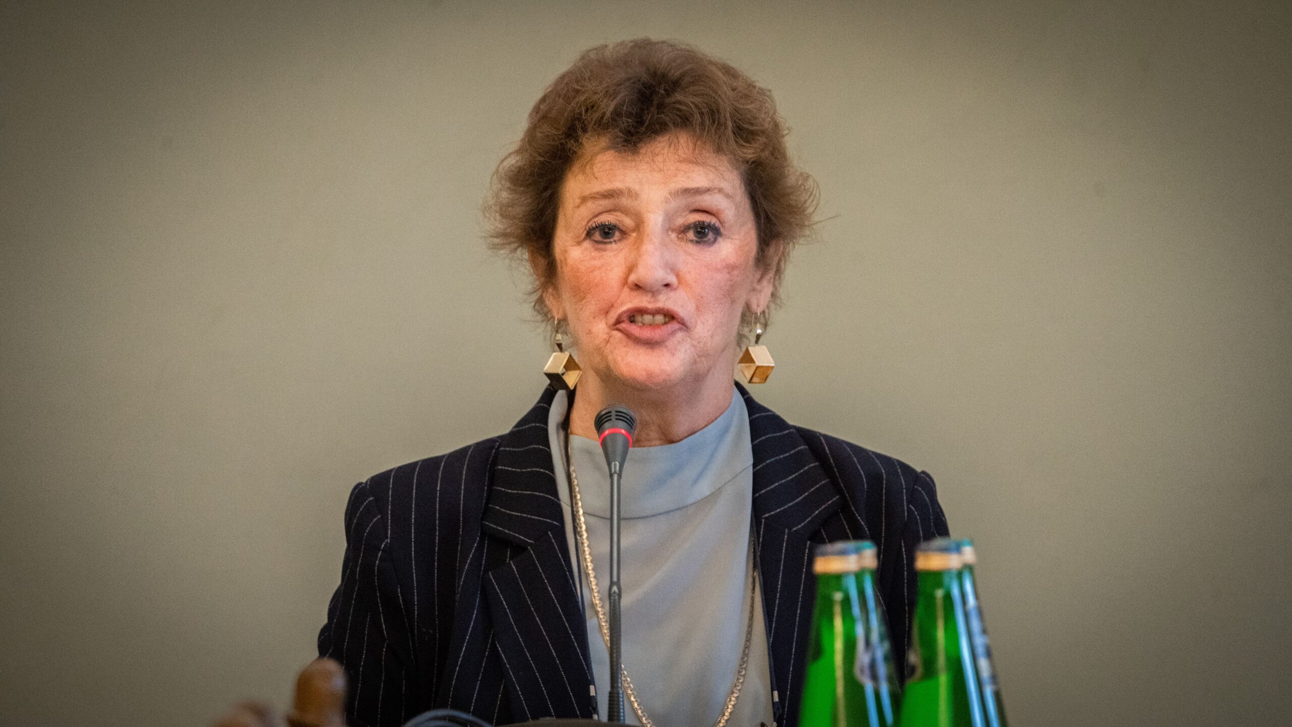 Kobieta w eleganckim stroju, z mikrofonem przed sobą, wygłasza przemówienie w sali konferencyjnej.